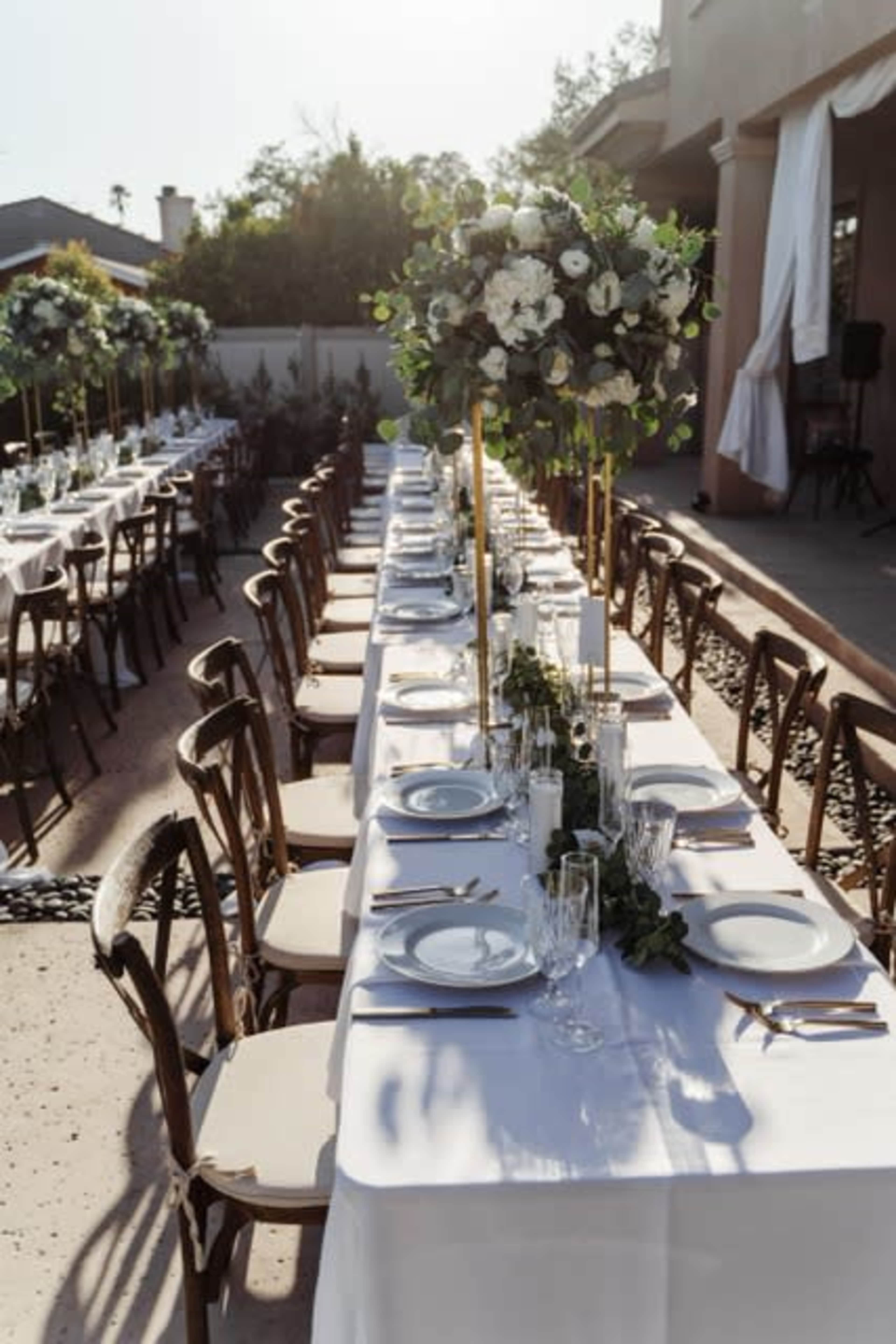 A long outdoor dining table is set with white linens, elegant tableware, and tall floral centerpieces under soft sunlight.