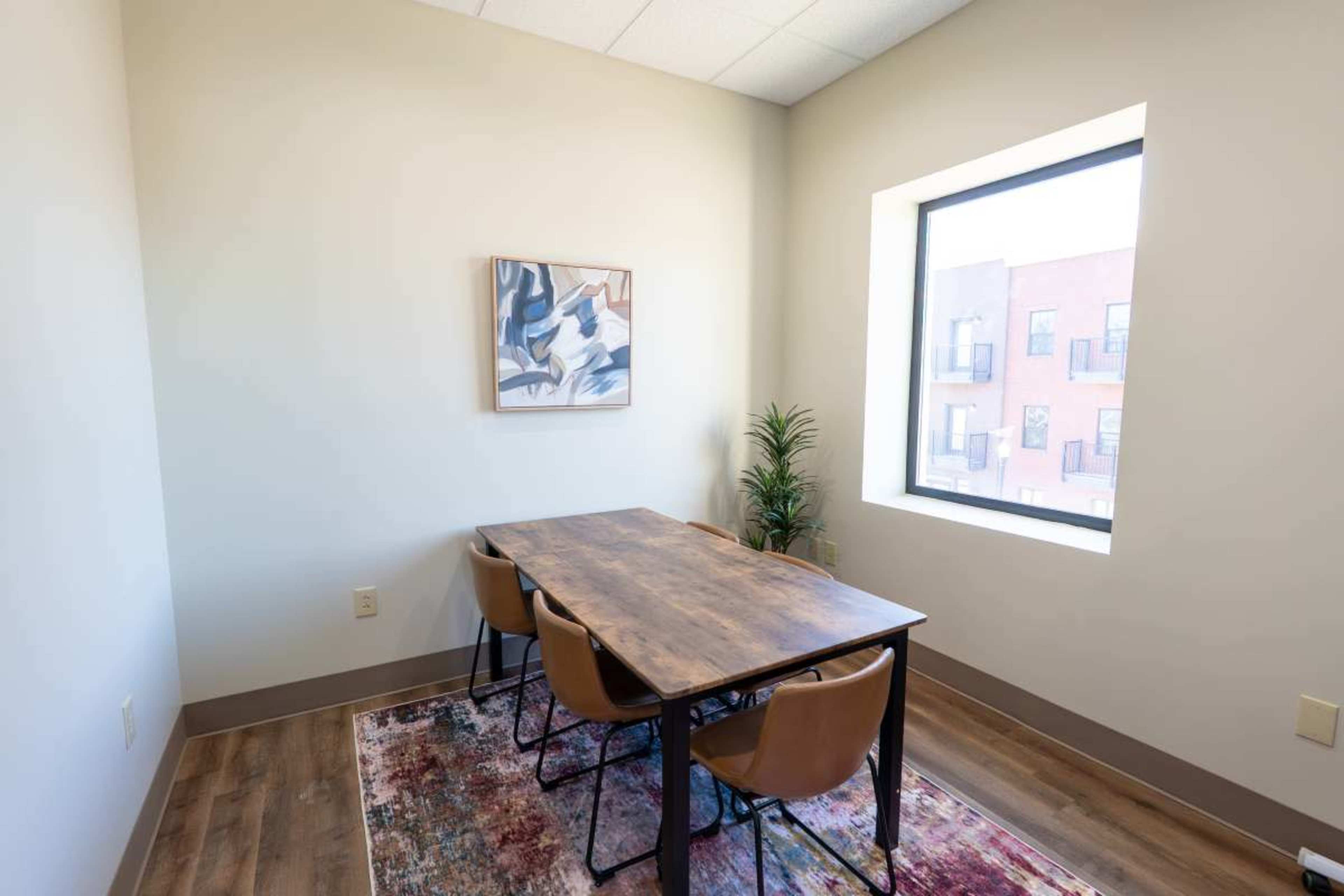 A small meeting room features a wooden table surrounded by four brown chairs, with a large window and a piece of wall art.