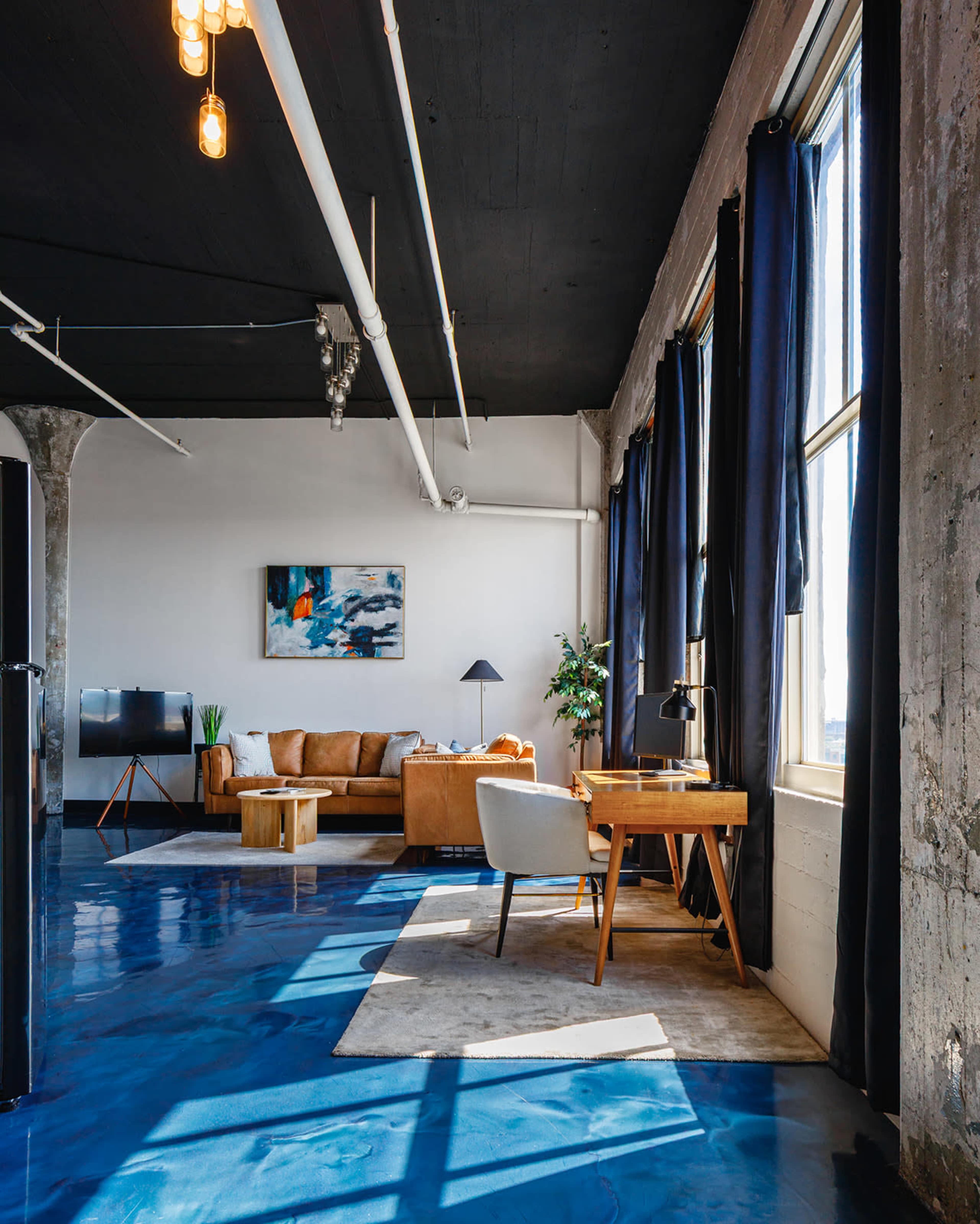The image shows a spacious, modern living area with a leather sofa, a wooden coffee table, a desk with a lamp, and large windows dressed with dark curtains, all set against a polished blue floor.