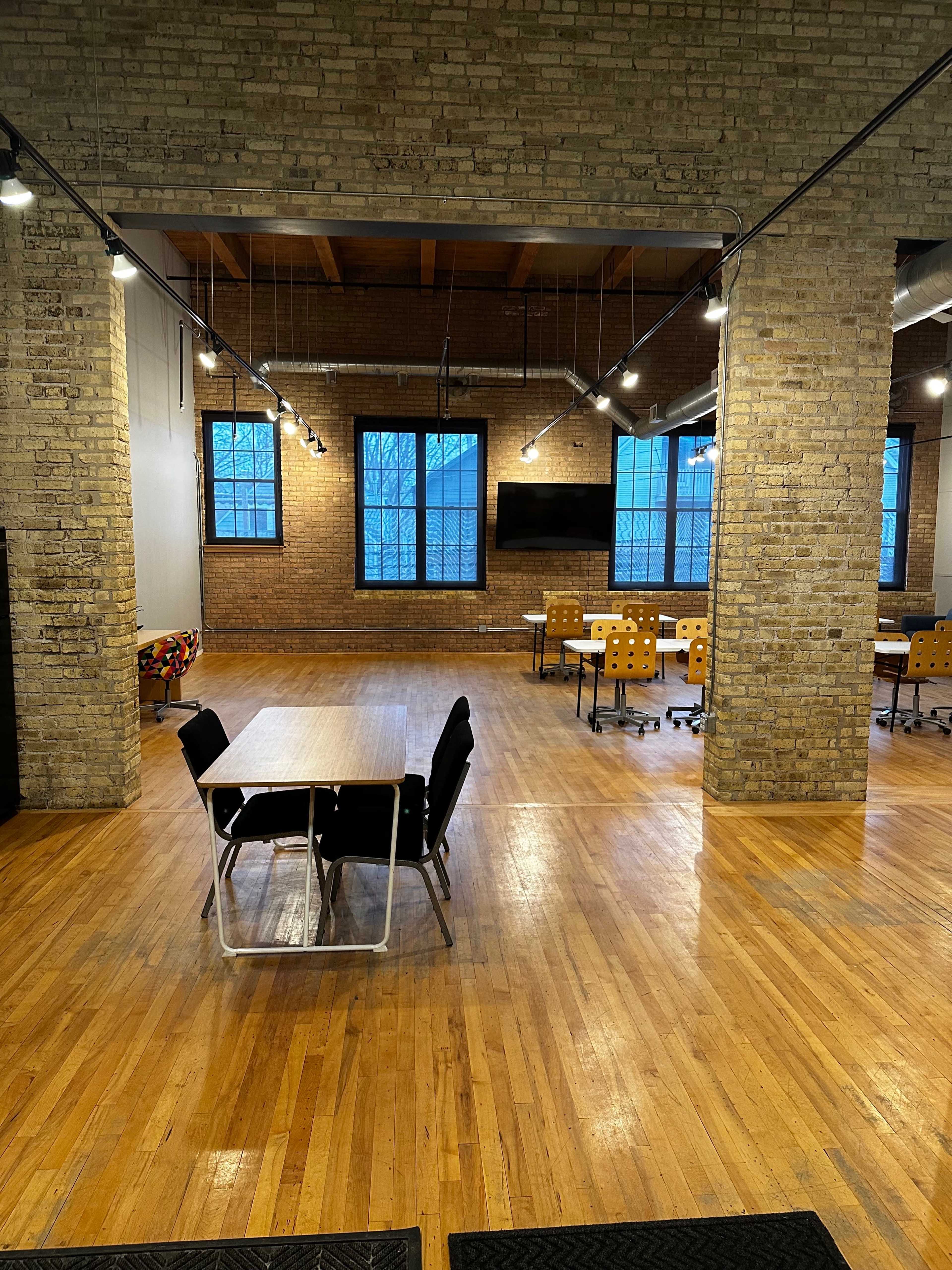 A spacious office area with wooden floors, brick walls, multiple tables and chairs, and large windows allowing natural light.