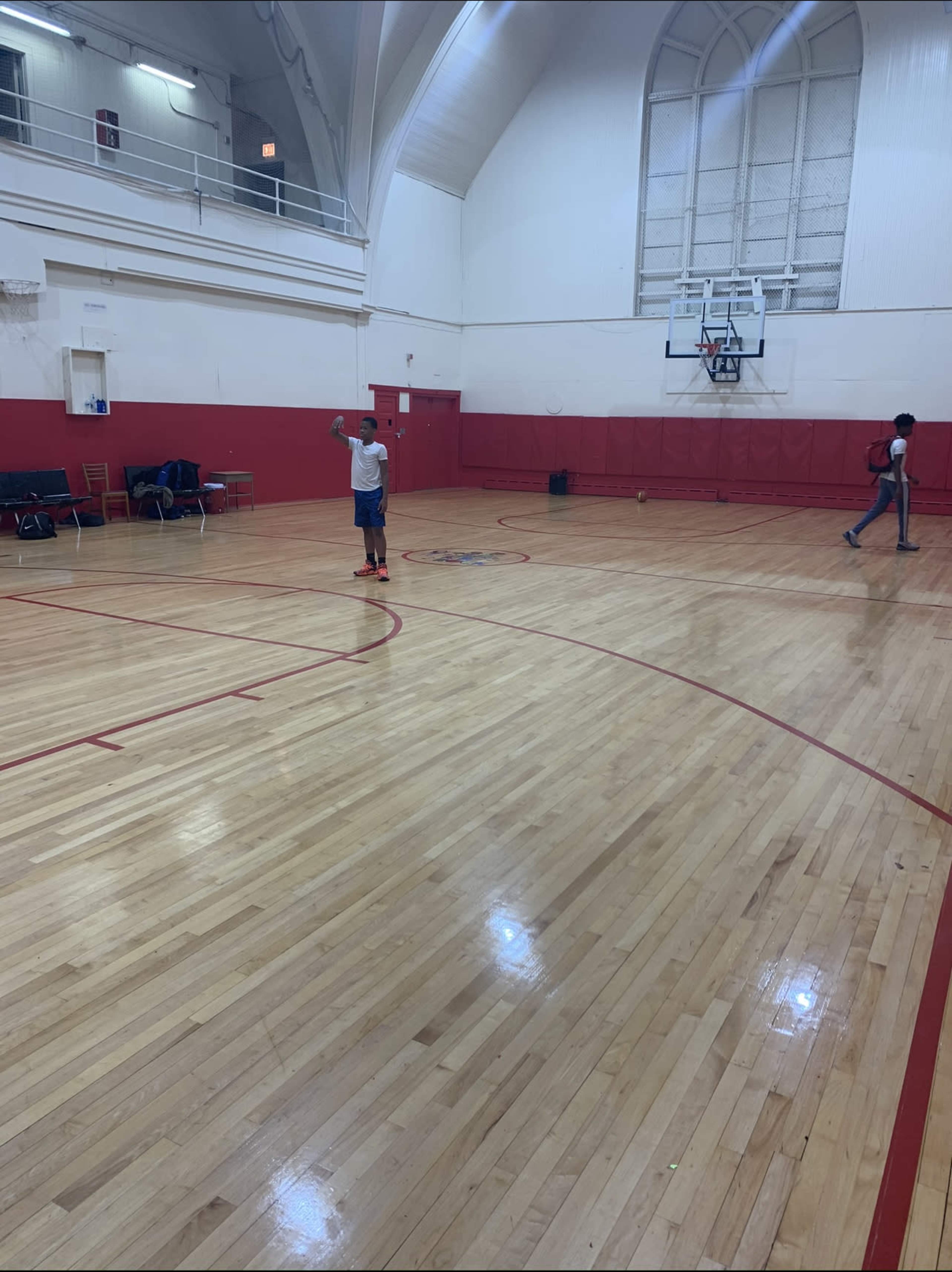 A lone person stands in the center of an empty gymnasium with a basketball court, while another individual walks in the background.