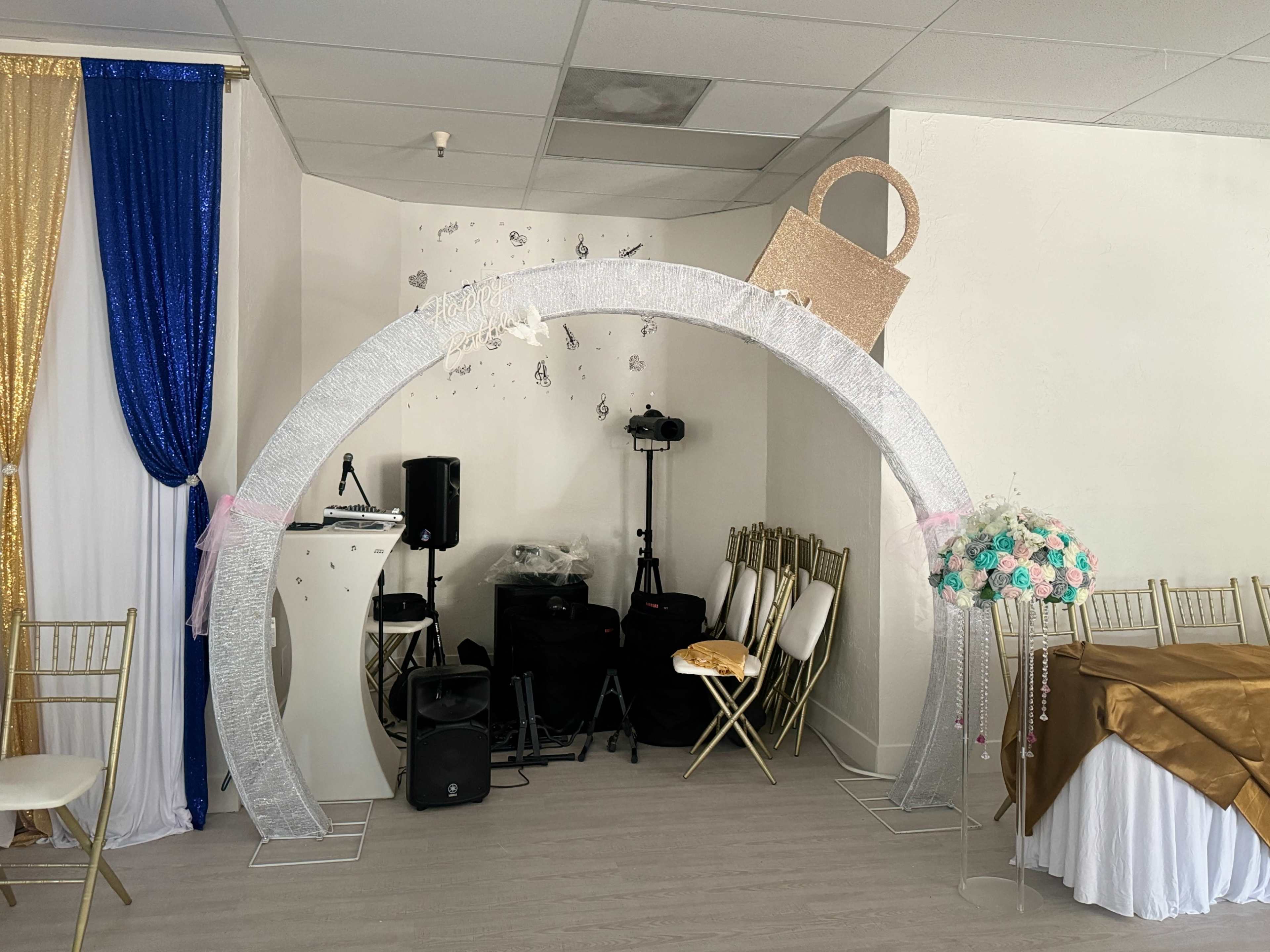 The image shows a decorative arch set up for an event, with a microphone and speakers in the background and a floral arrangement nearby.