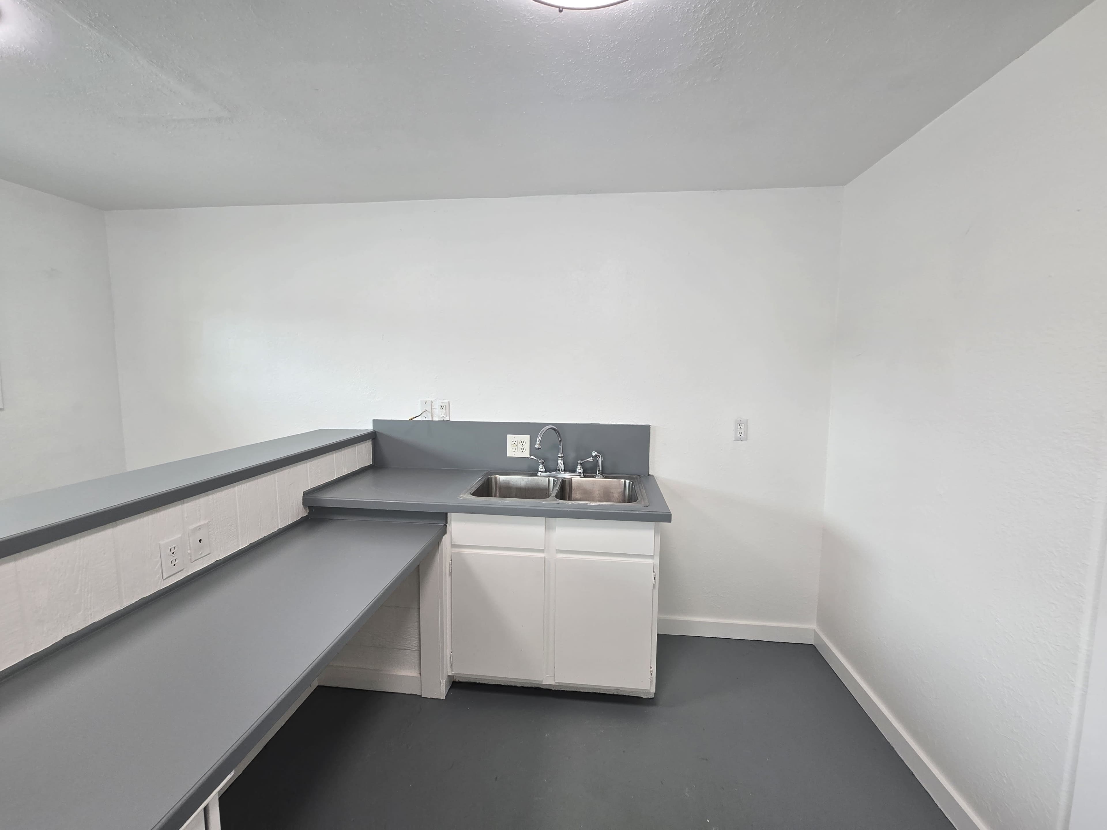 The image shows a minimalistic kitchen space with a gray countertop, a double sink, and white cabinetry against plain white walls.
