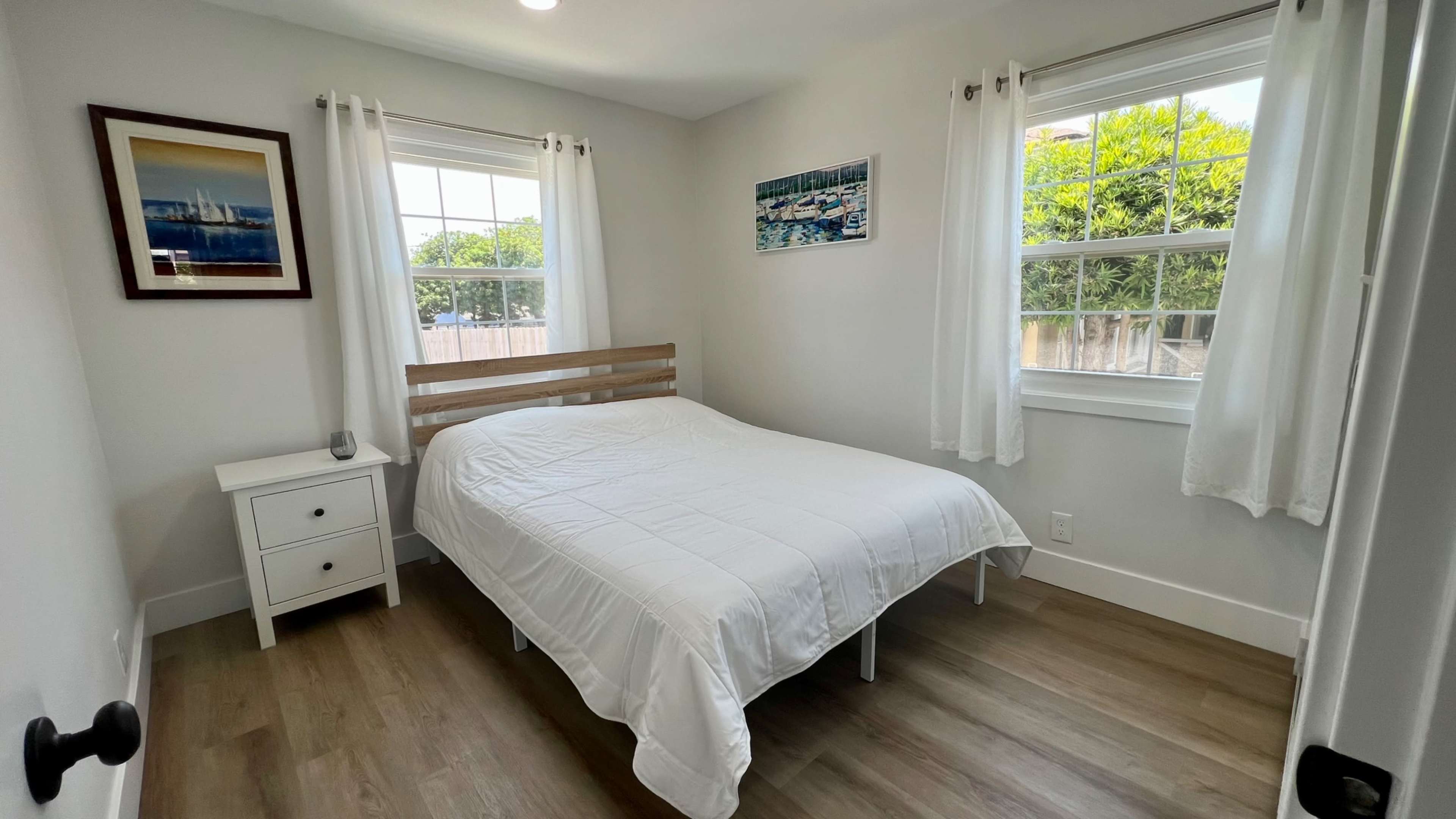 A bright bedroom featuring a bed with a white comforter, a nightstand, large windows, and light-colored walls.