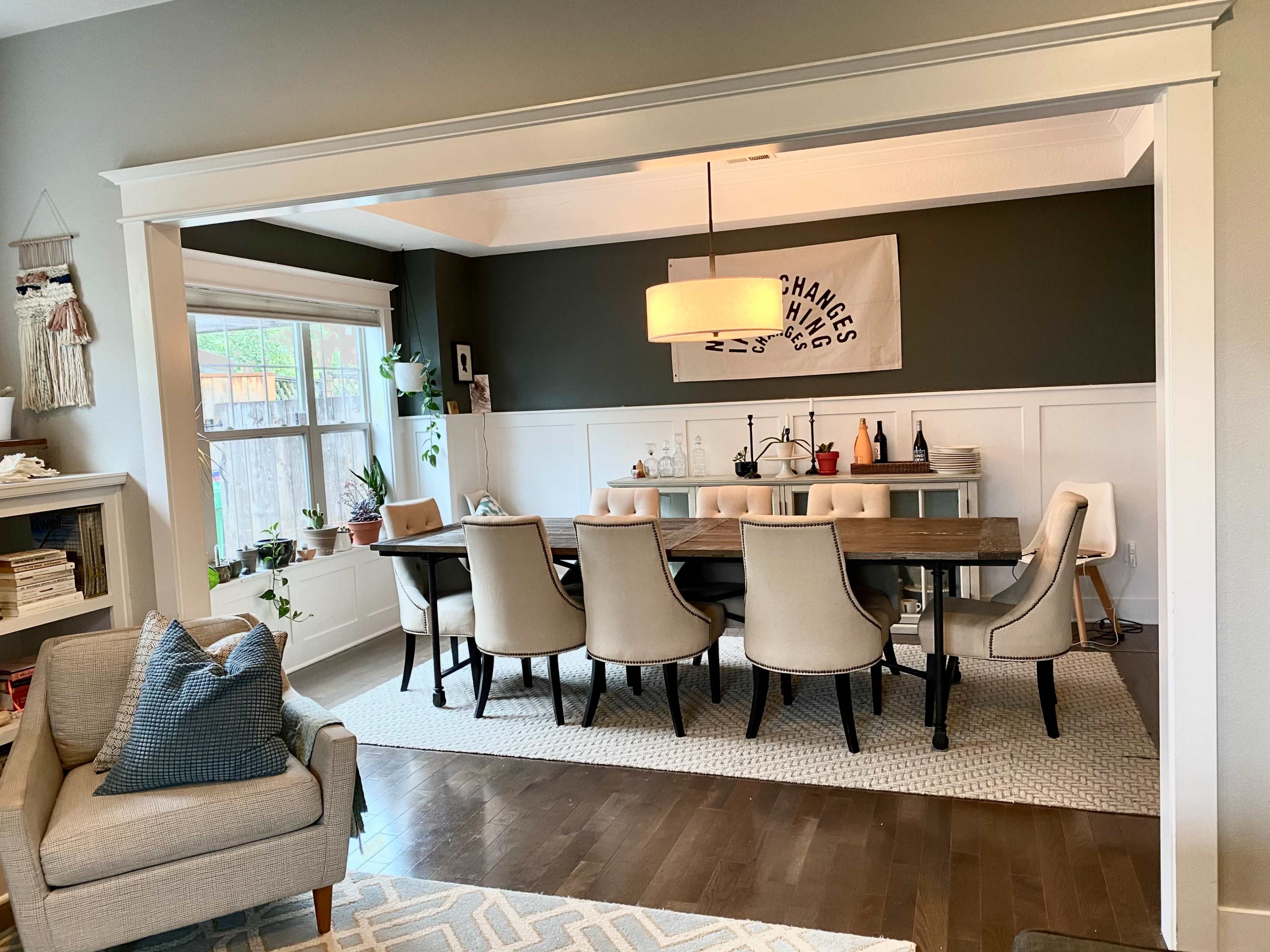 A dining area features a long table surrounded by beige chairs, with a modern light fixture above and decorative items on a side table.