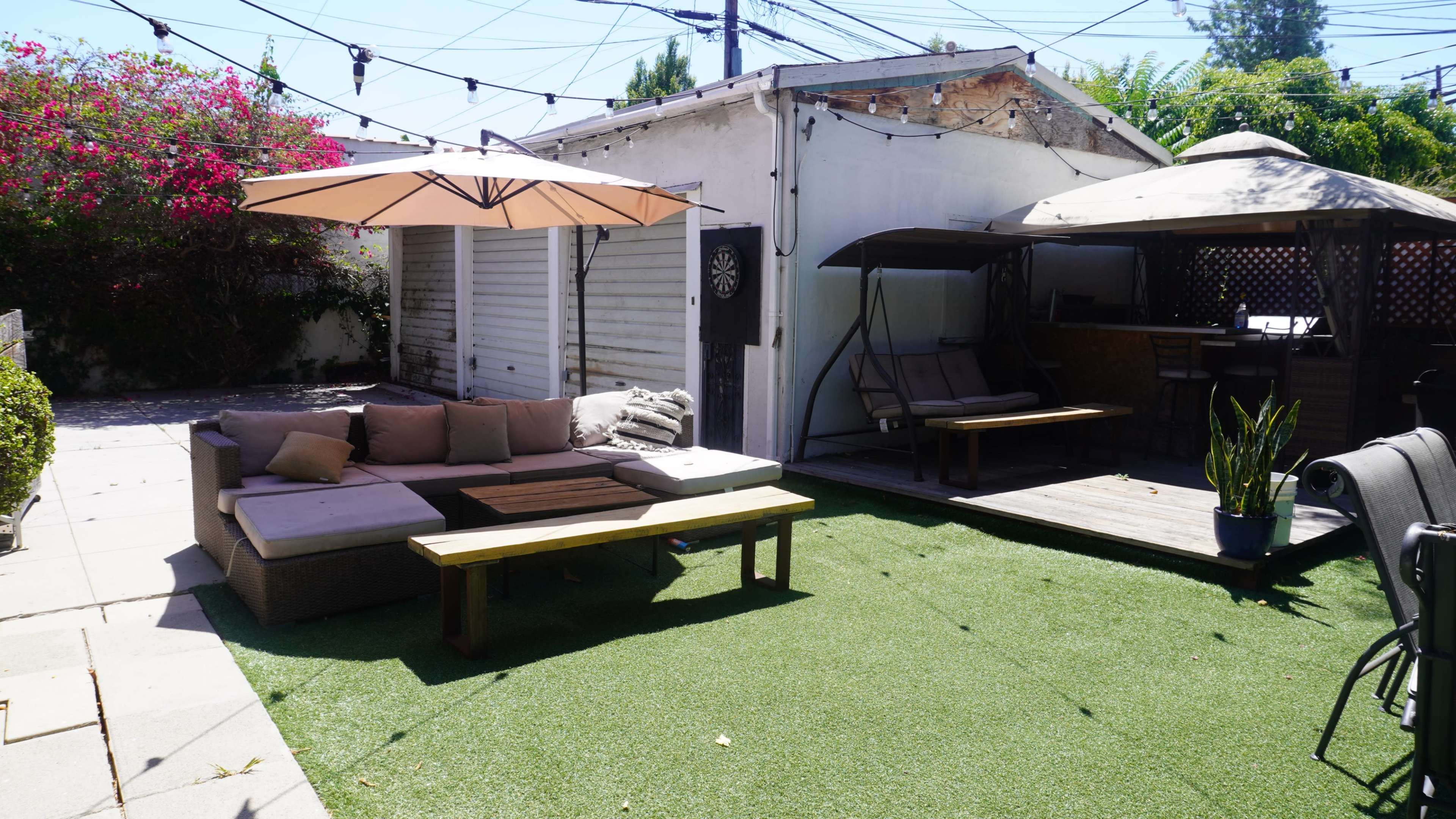 The scene features a patio area with a sectional sofa, a wooden coffee table, two shaded lounge areas, and artificial grass surrounded by vibrant plants and utility lines.