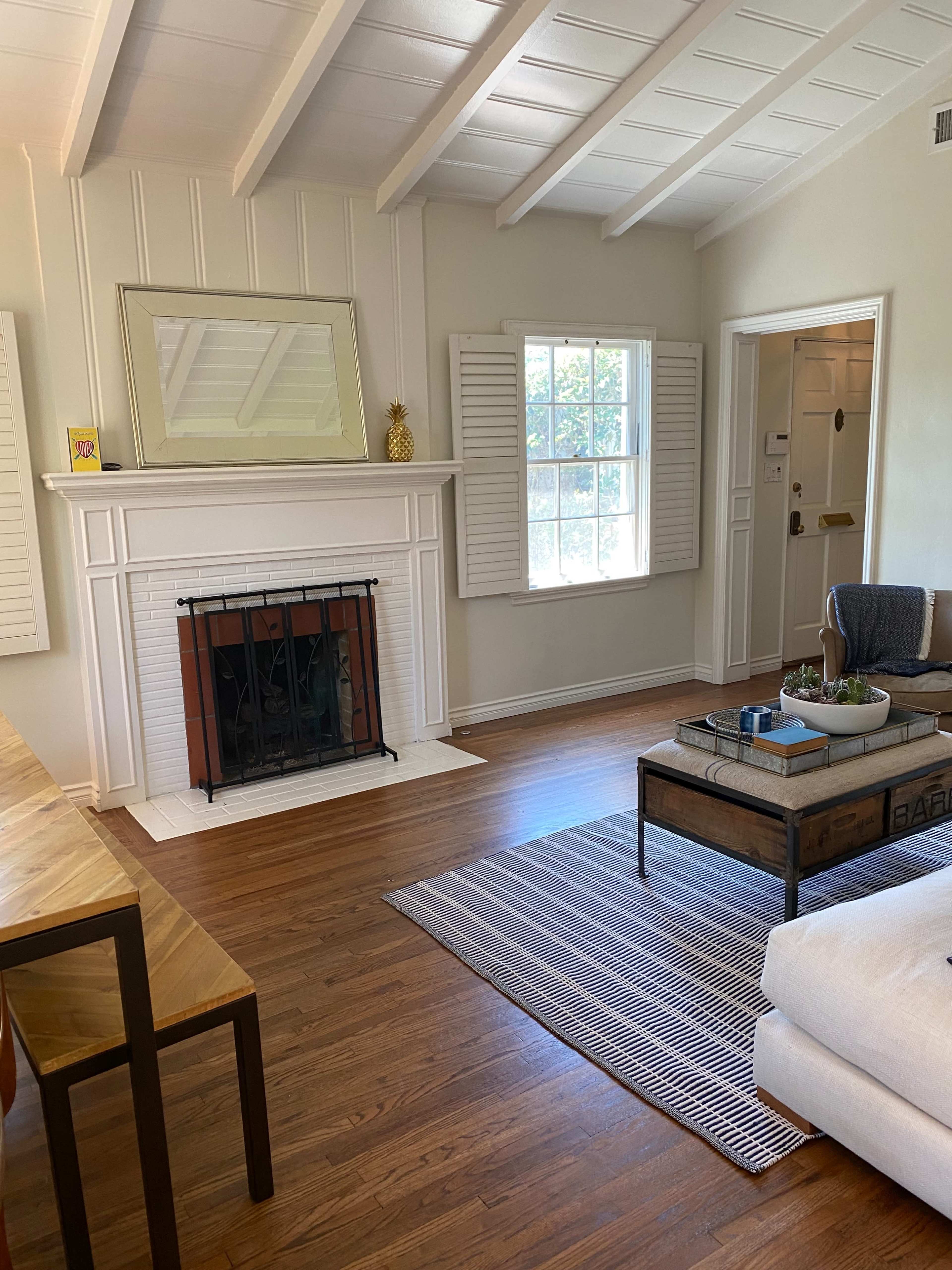 A spacious living room features a white fireplace, a large mirror, and a window with shutters, accompanied by a coffee table and a light-colored sofa.