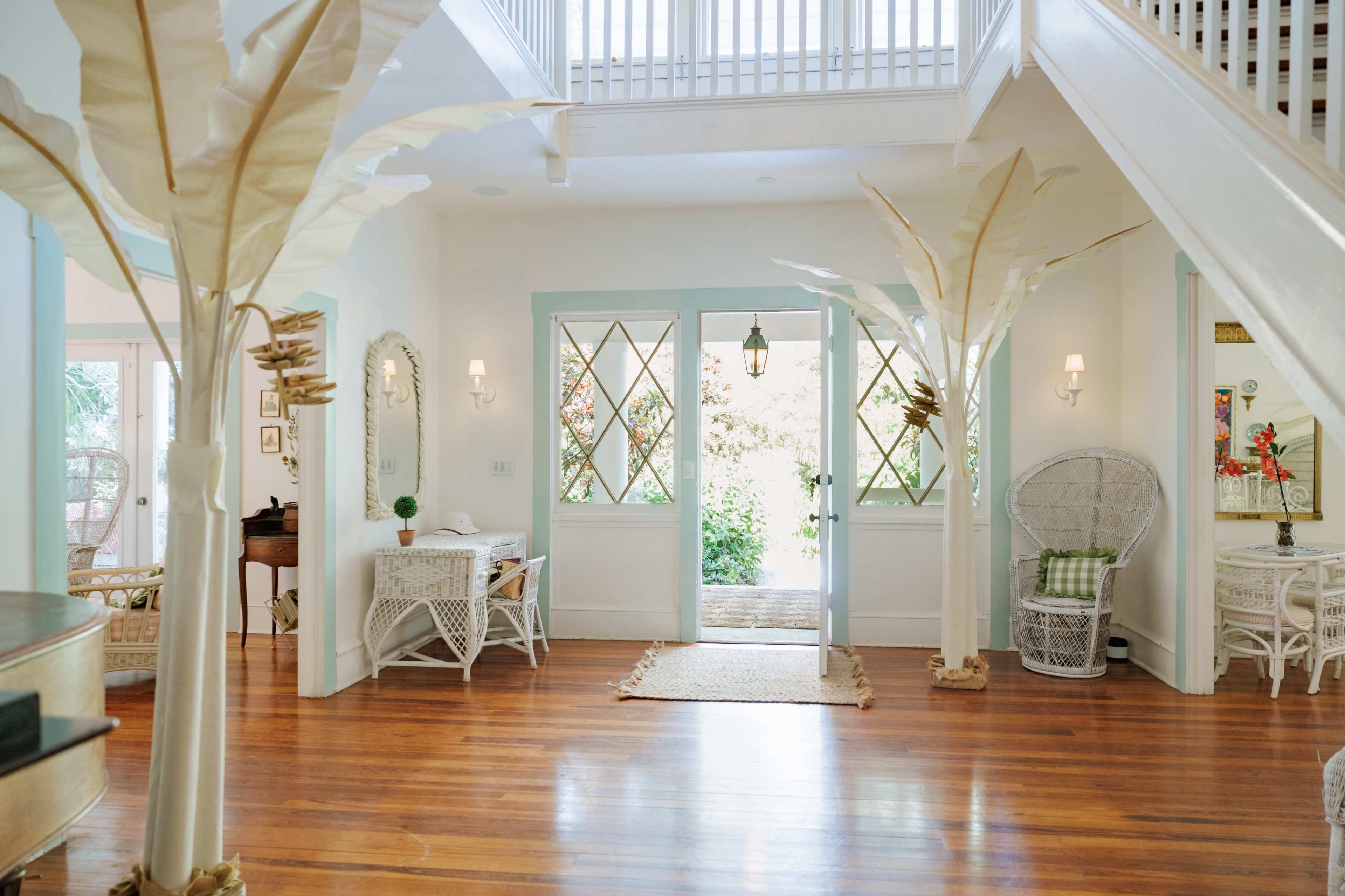 The image shows a bright entryway featuring white walls, wooden floors, and decorative palm-like plants flanking a door with diamond-shaped windows.