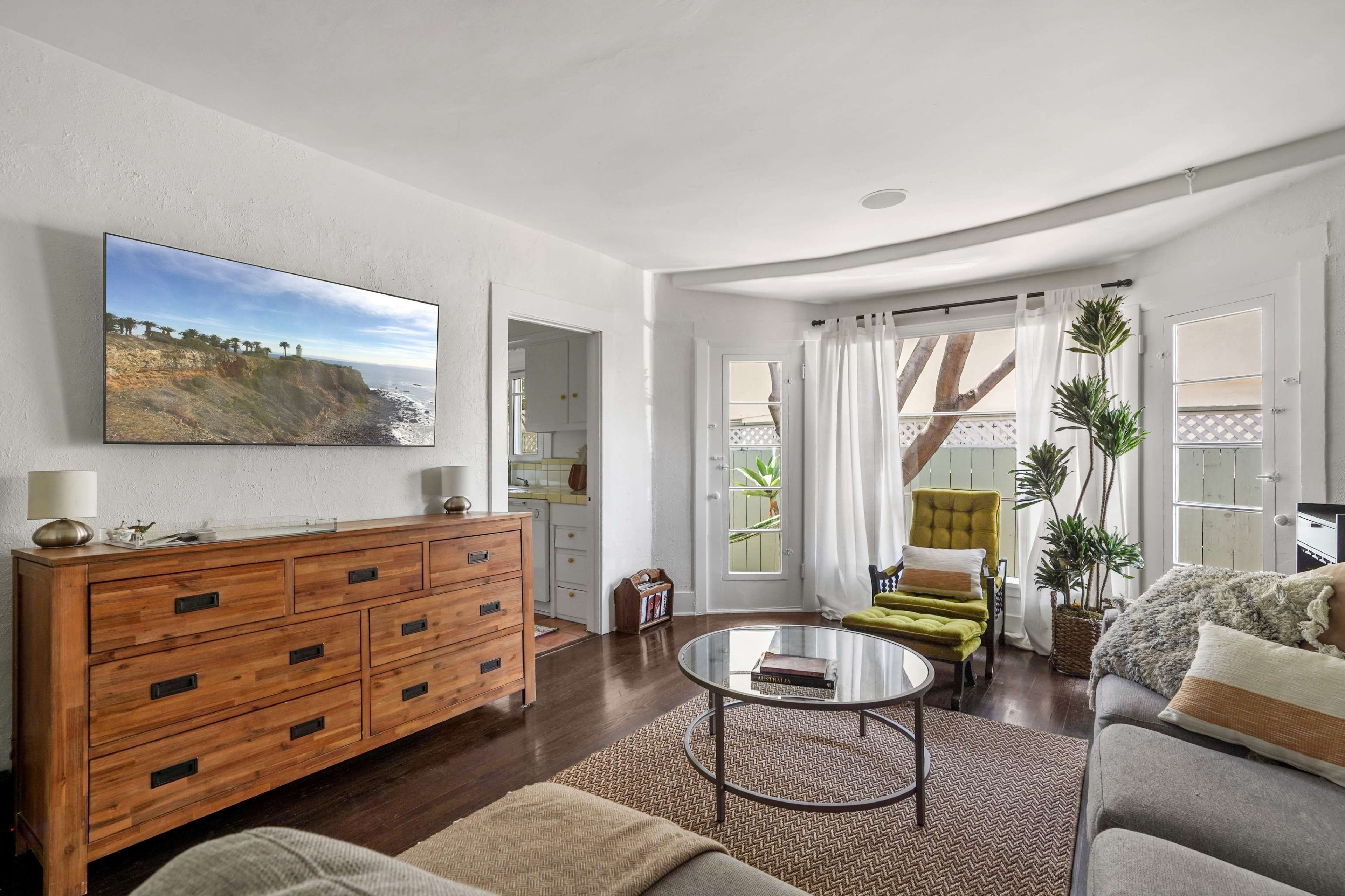 The living room features a television mounted on a white wall, a wooden dresser, a round coffee table, and a green accent chair, all arranged around large windows that allow natural light in.