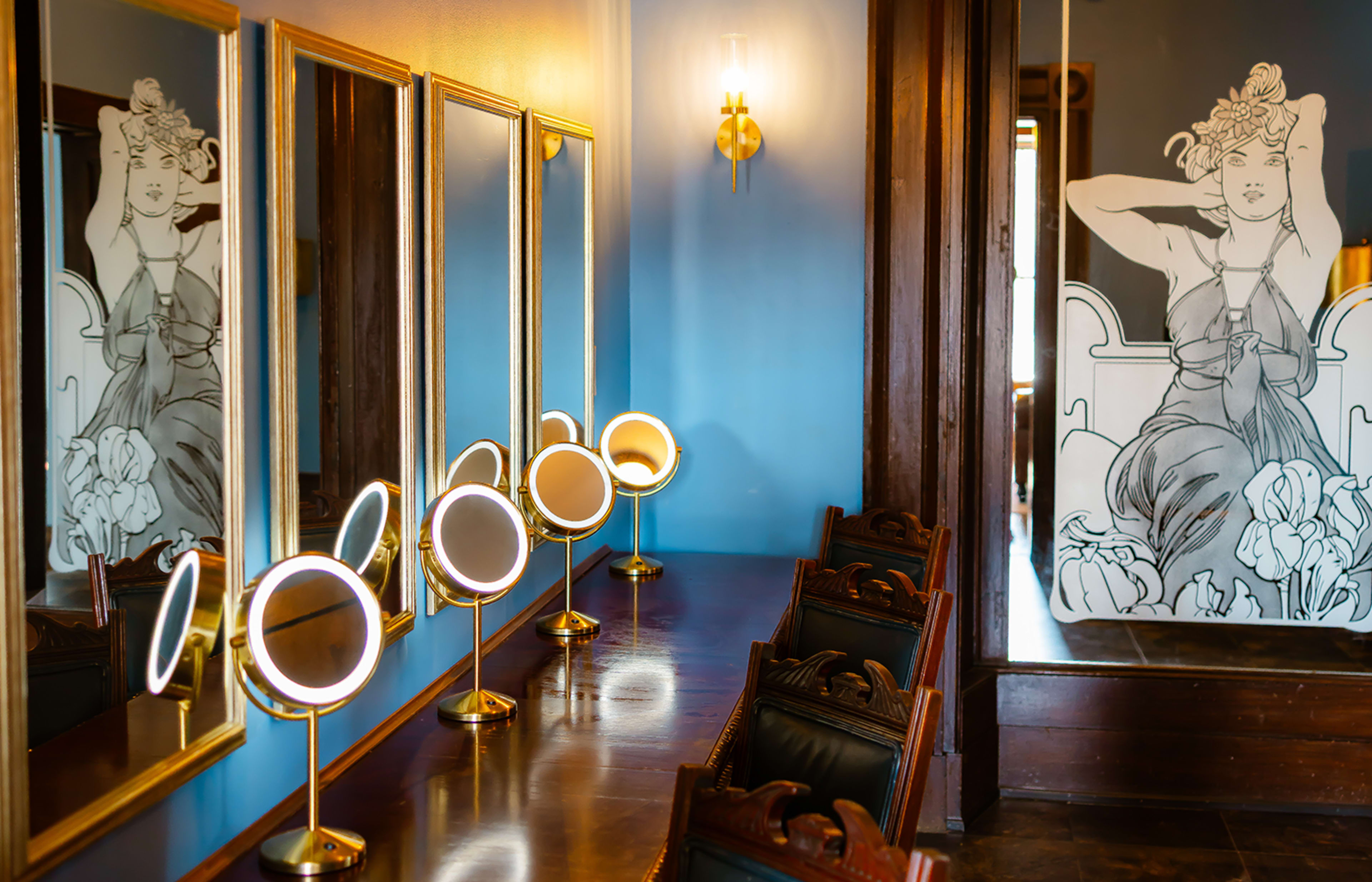 The image shows a makeup vanity area with multiple illuminated mirrors and a large decorative mural of a seated woman on a blue wall.