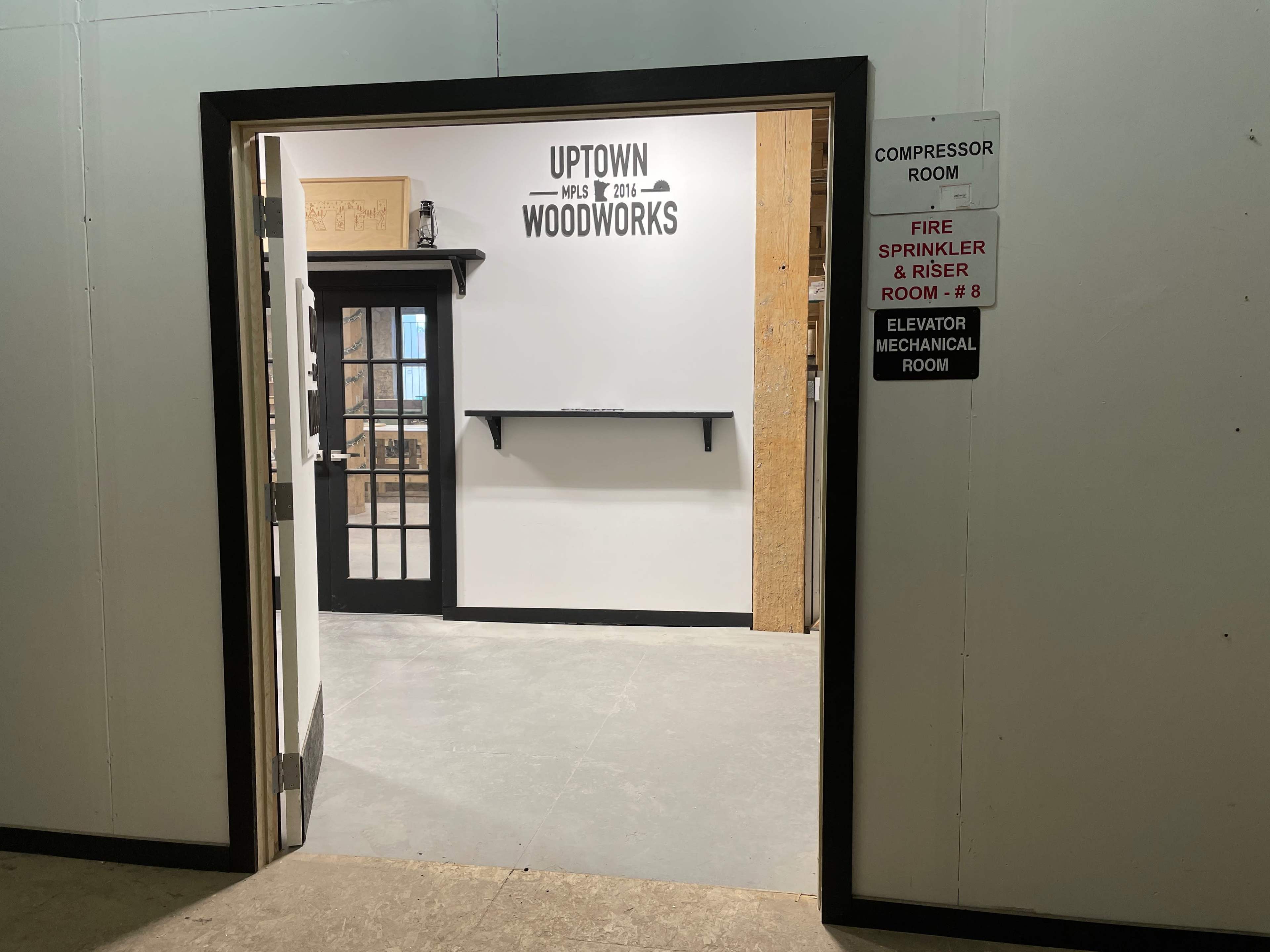 The image shows an open doorway leading into a workshop area with a sign reading "Uptown Woodworks" on the wall and a shelf mounted beside the entrance.