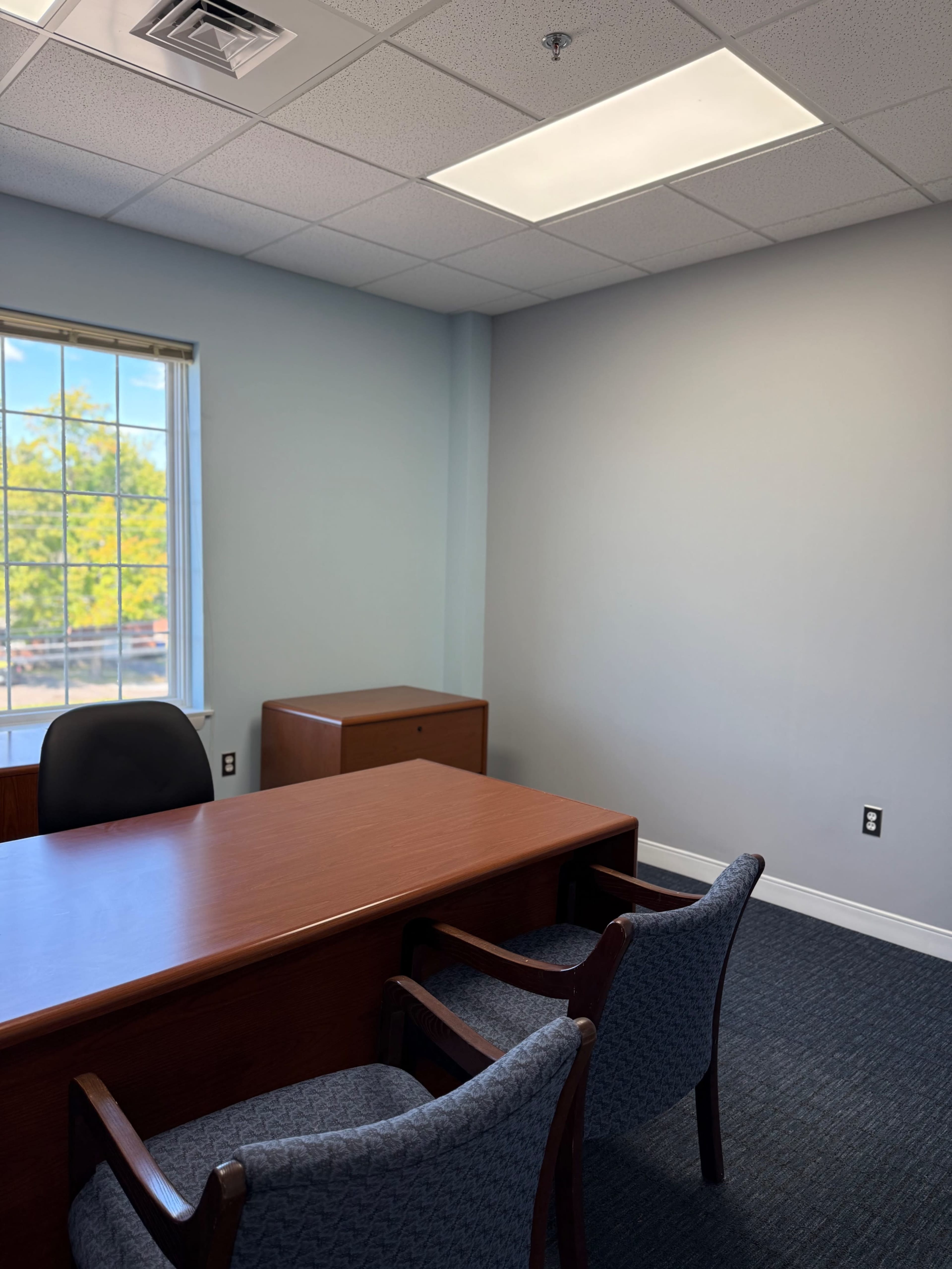 The image shows a small office with a wooden desk, a chair, and a window providing natural light from outside.