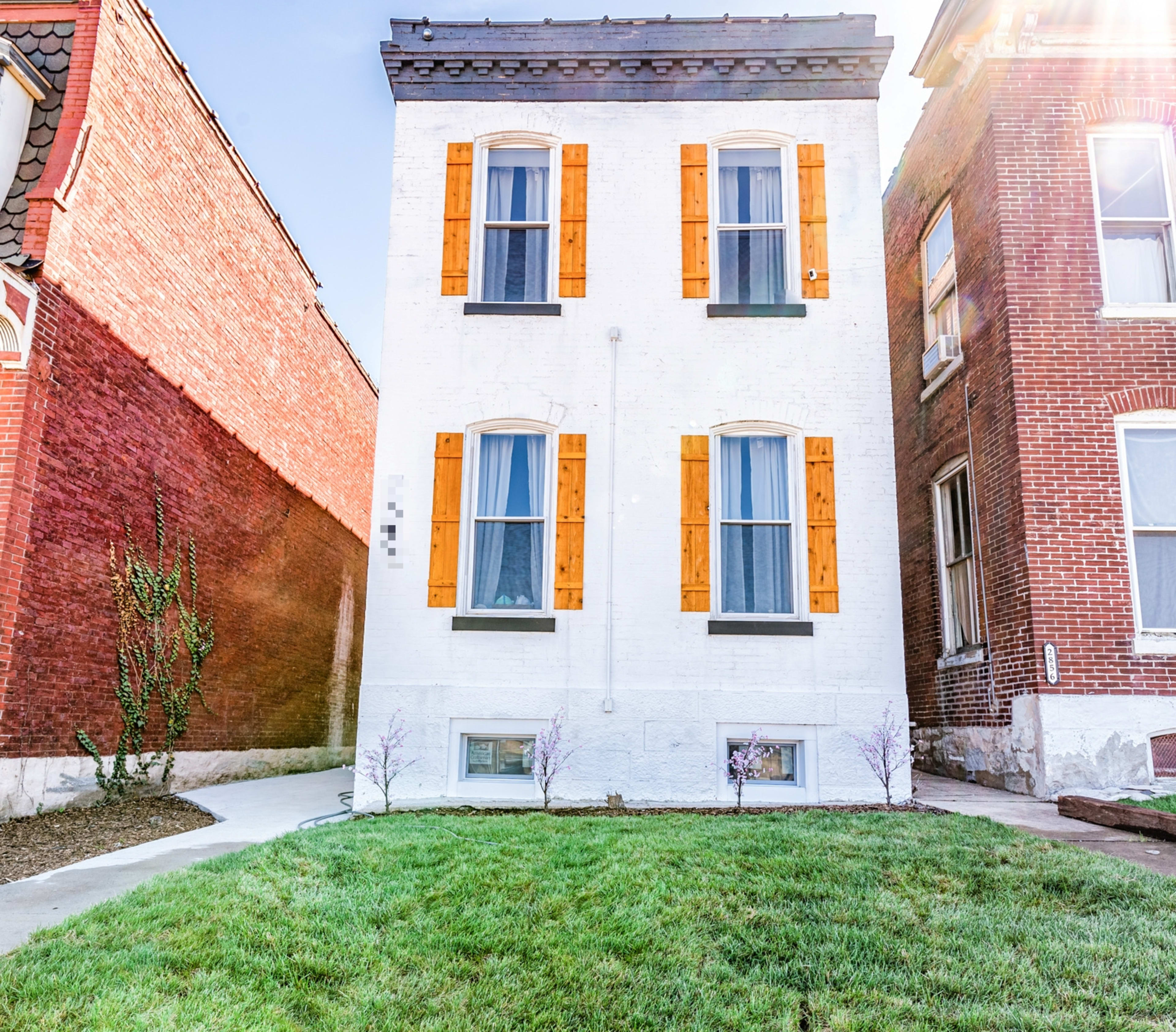 A two-story white house with orange shutters and large windows, situated between two brick buildings on a grassy area.