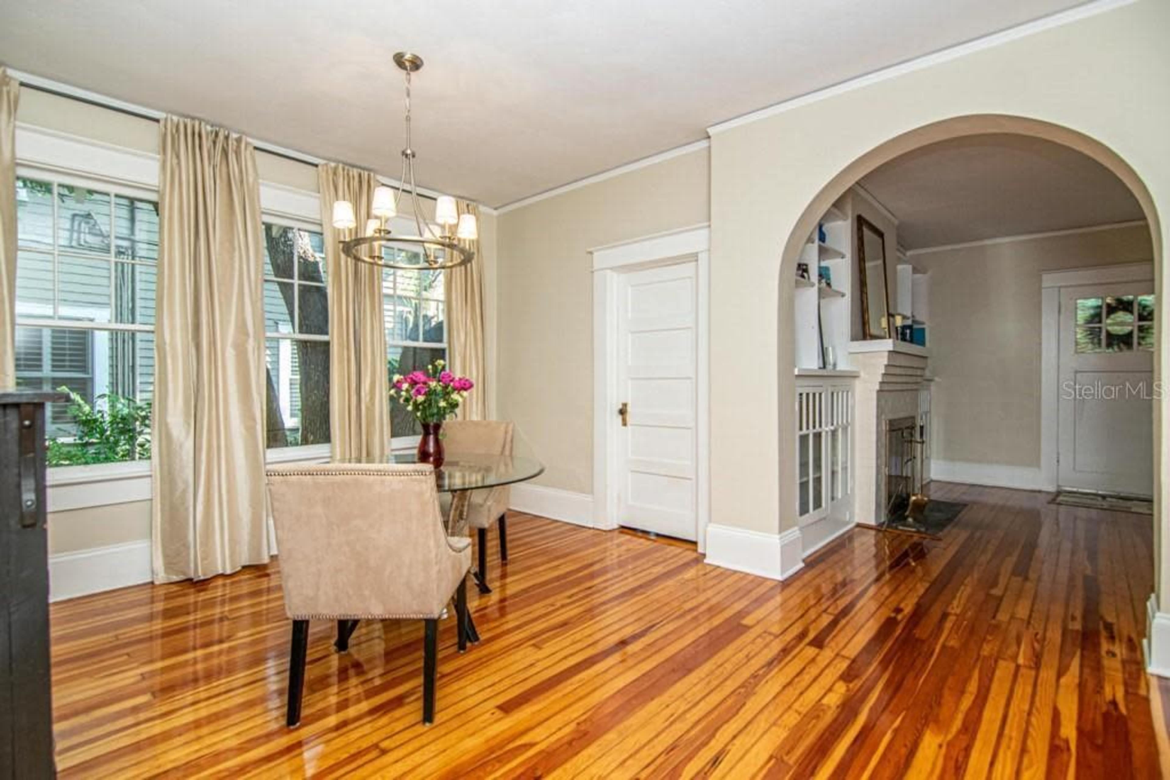 The image shows a dining area featuring a round glass table with two chairs, hardwood flooring, and large windows with curtains.