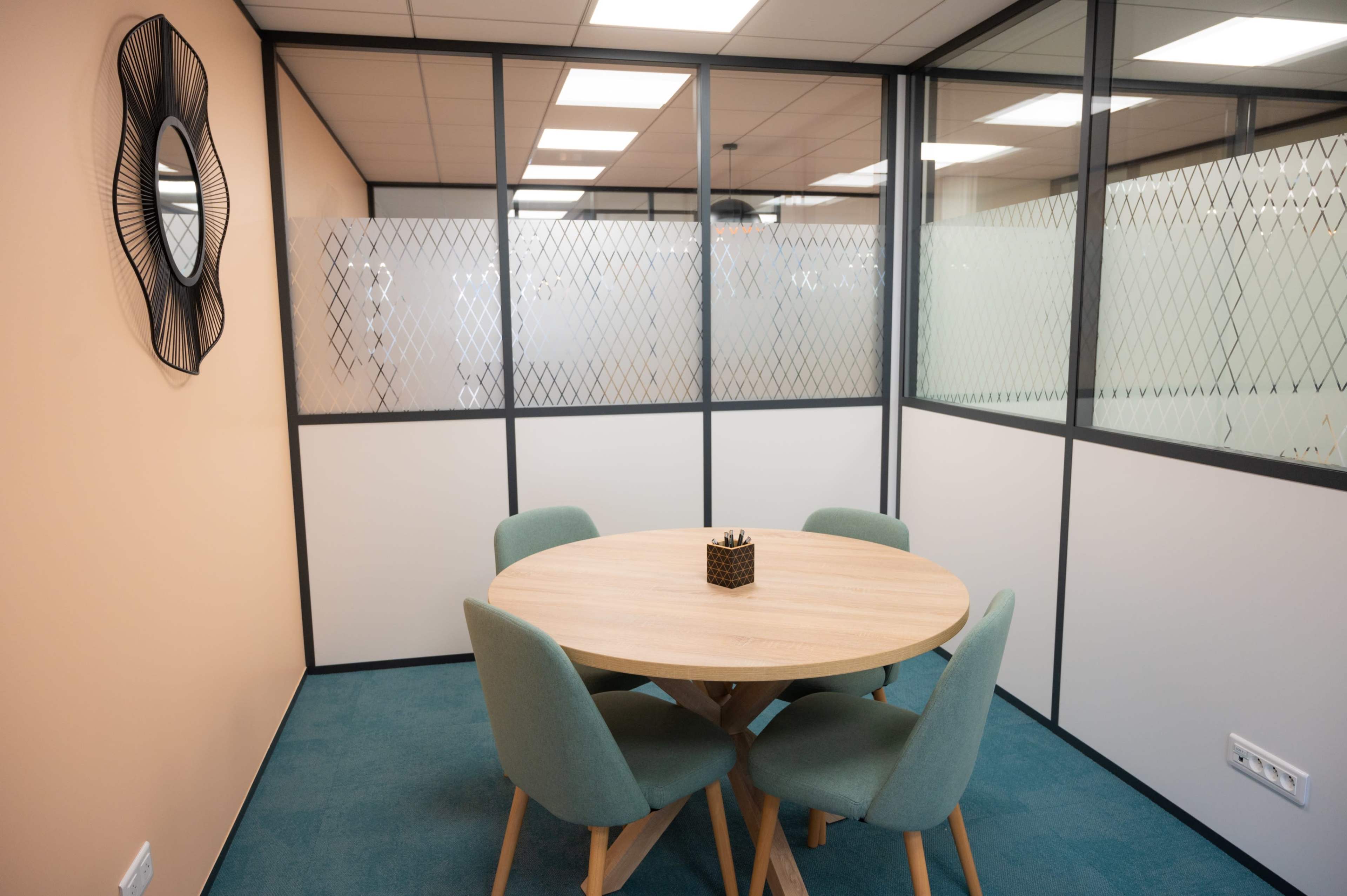 The image shows a modern meeting room with a round wooden table surrounded by four teal chairs, enclosed by glass walls.