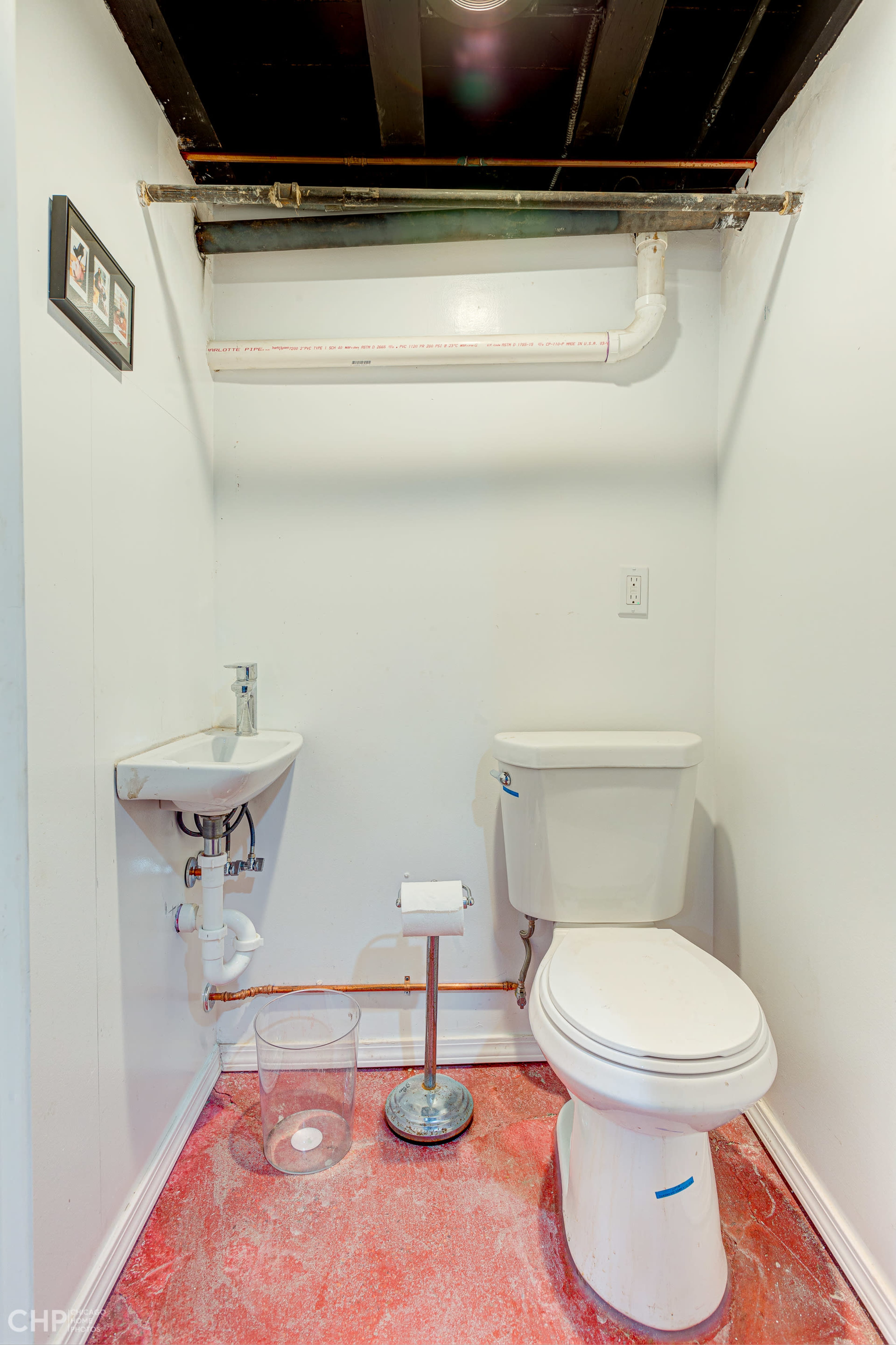 A small, unfinished bathroom with a toilet, a sink, and exposed plumbing on the walls and ceiling.