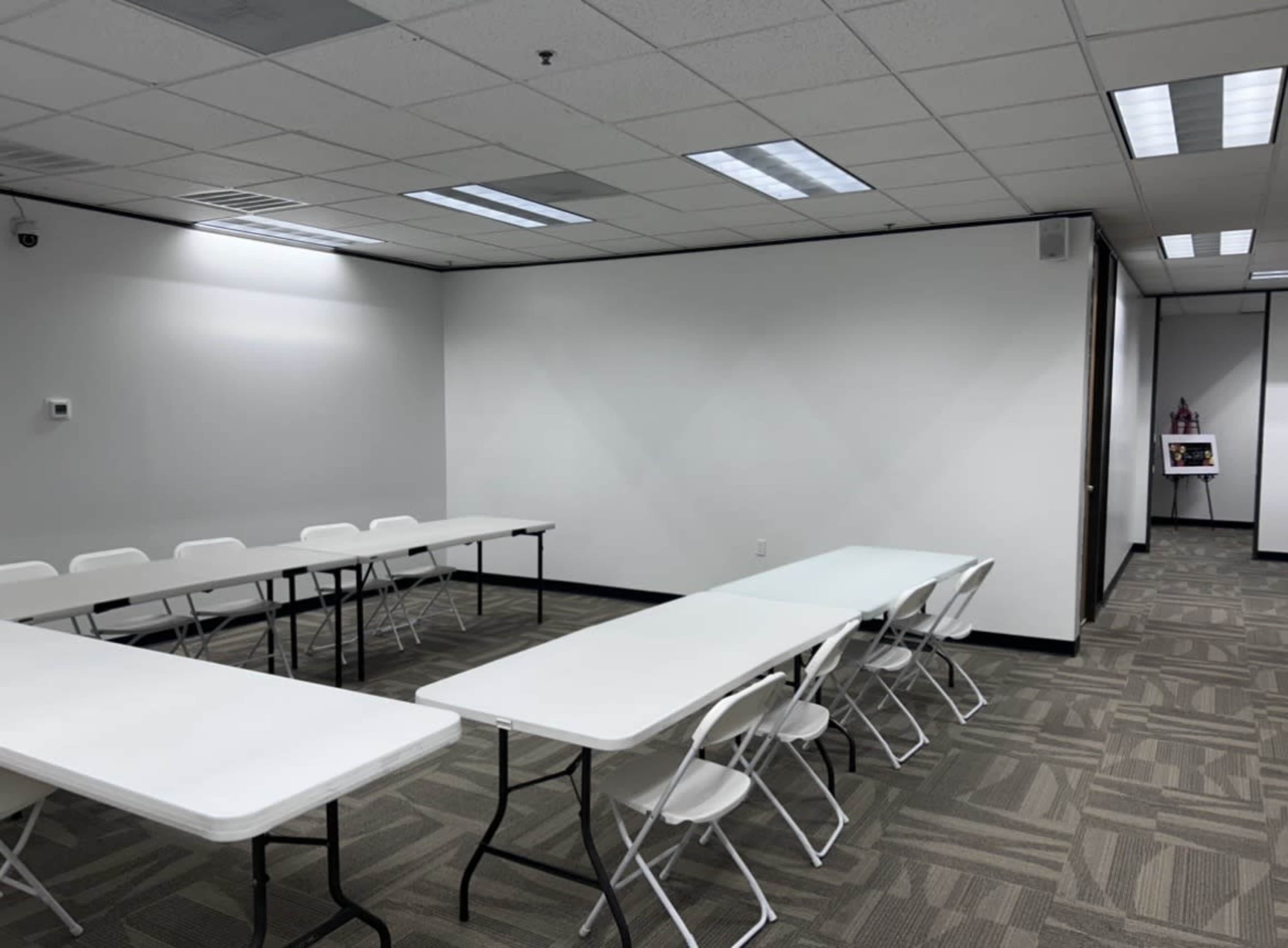 The image shows a spacious, empty meeting room with several rows of white folding tables and chairs arranged neatly, and a small display stand in the background.