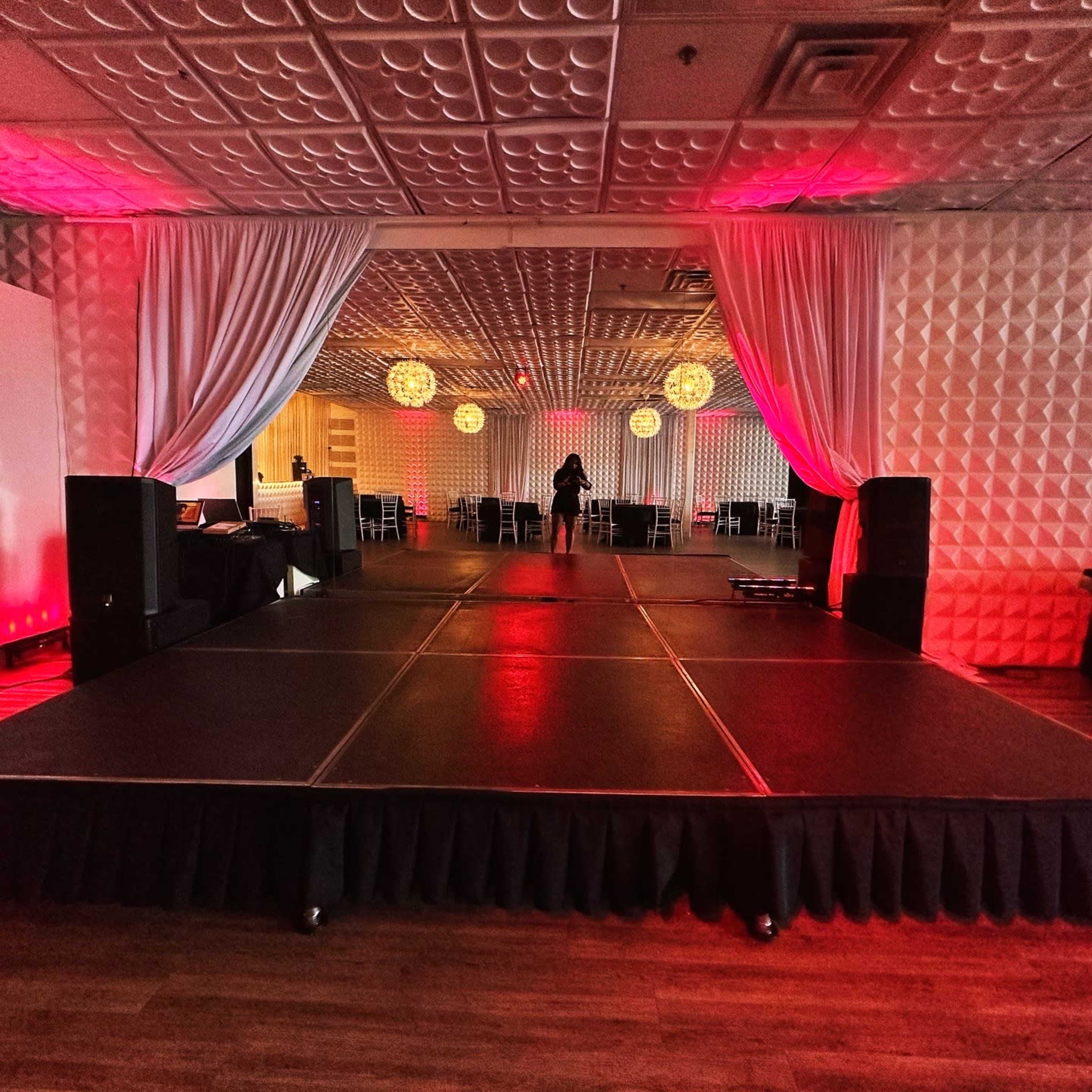 A stage is set in a warmly lit venue with draped curtains and decorative hanging lights.