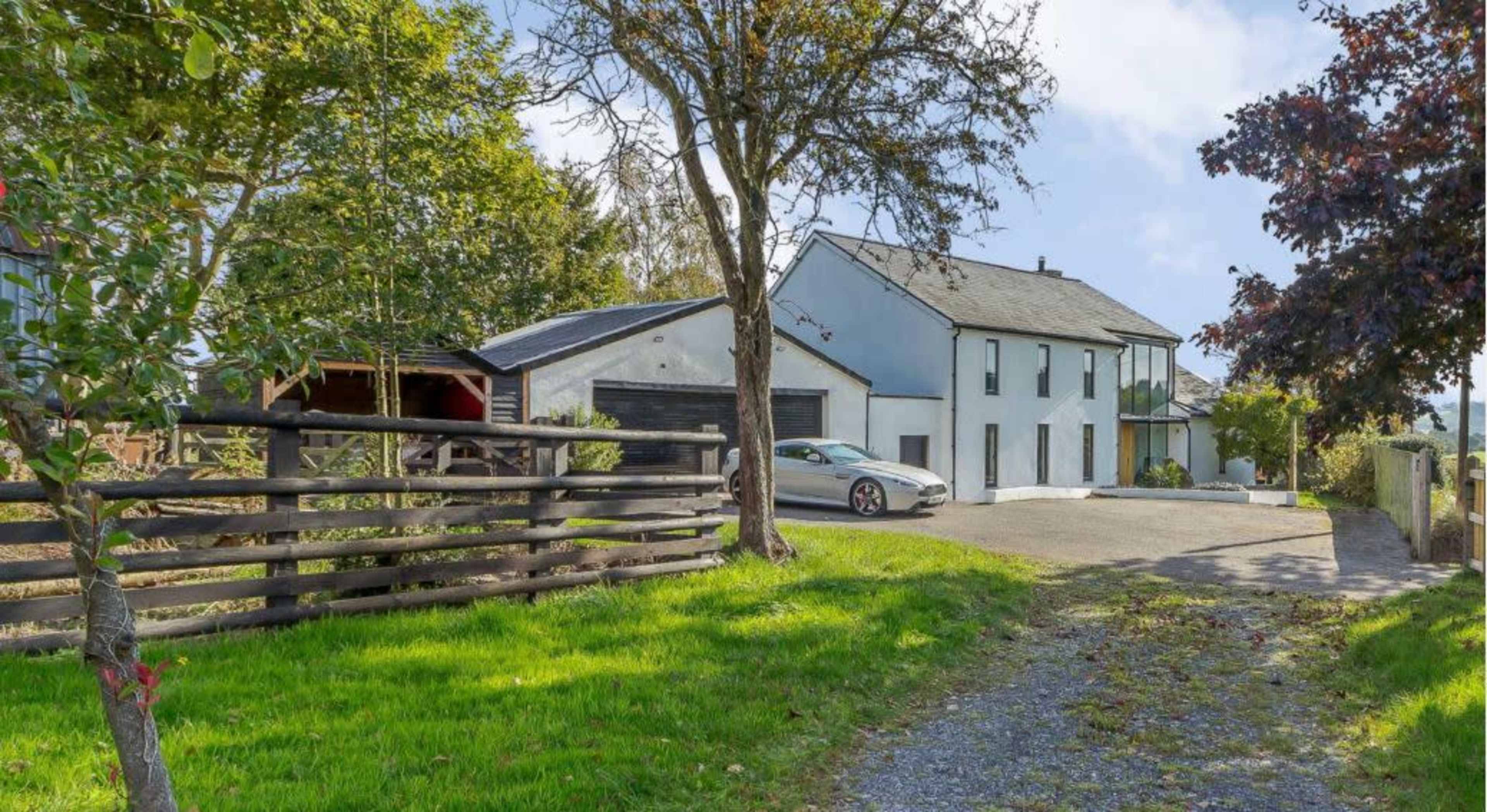 A modern two-story house with large windows sits beside a gravel driveway, surrounded by trees and a wooden fence.