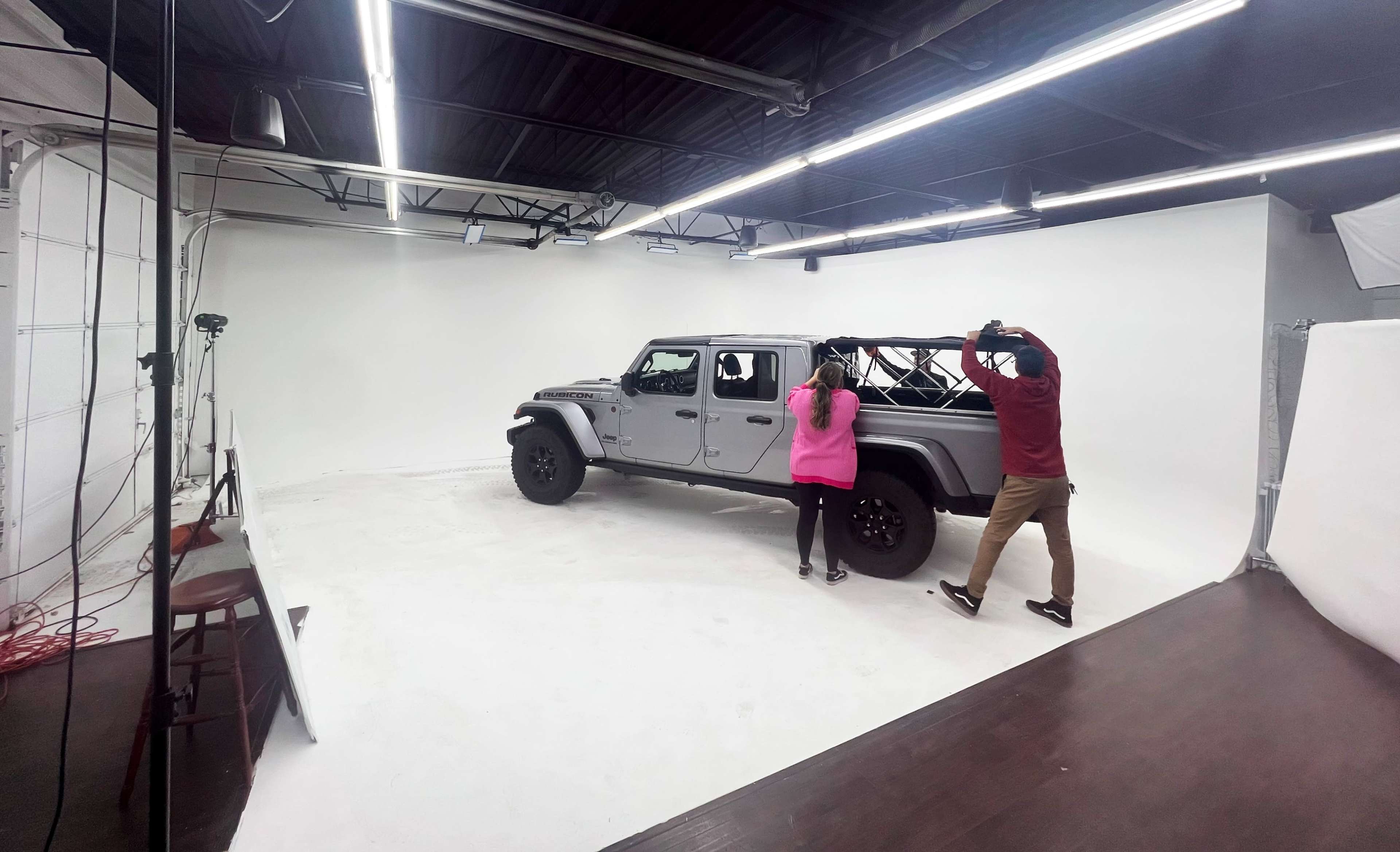 Two people are positioned near a parked Jeep Wrangler in a well-lit photo studio with a white backdrop.