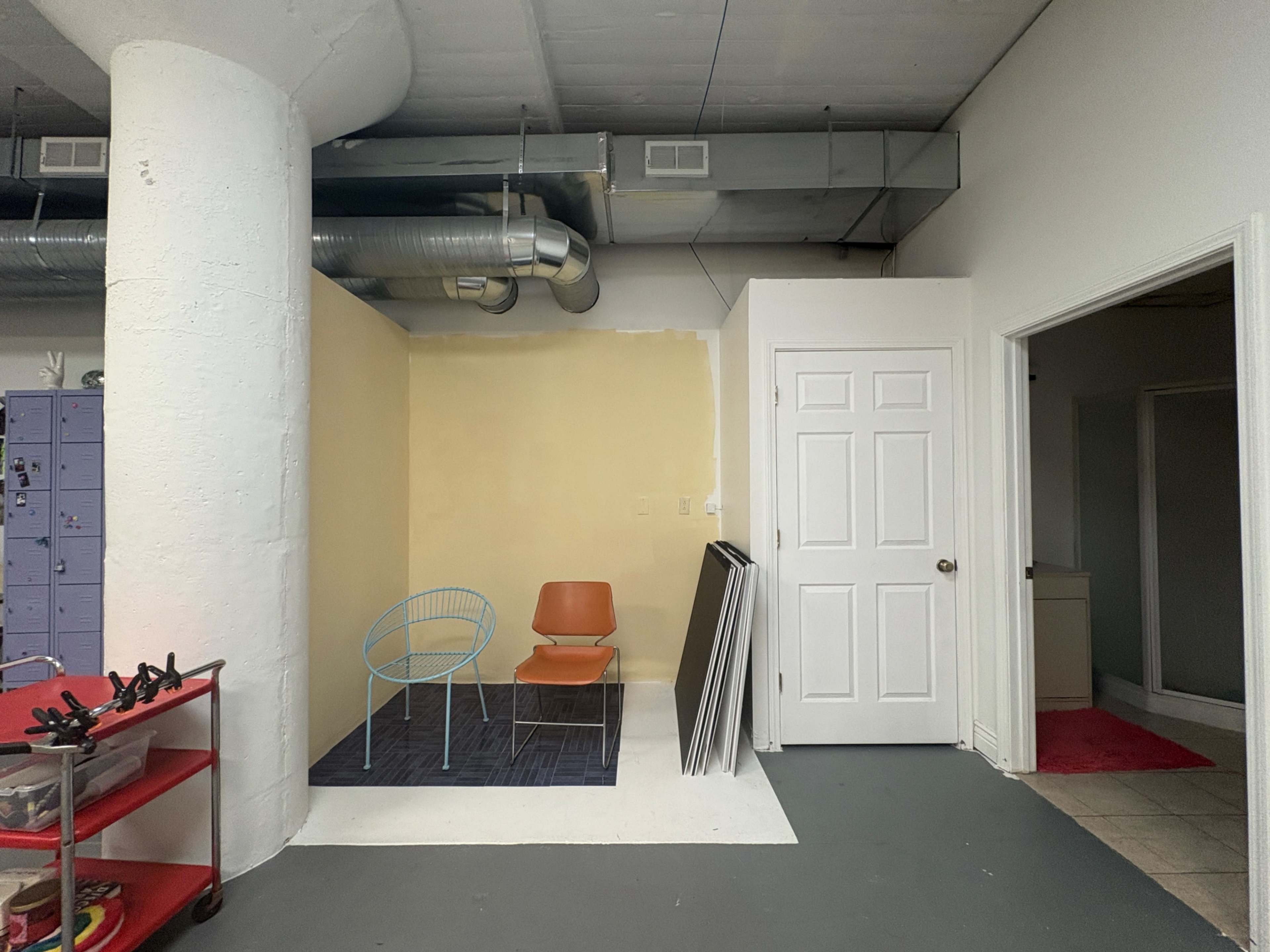 The image shows a spacious, partially painted room with a blue wire chair and an orange chair, a white door, and exposed ceiling ducts.