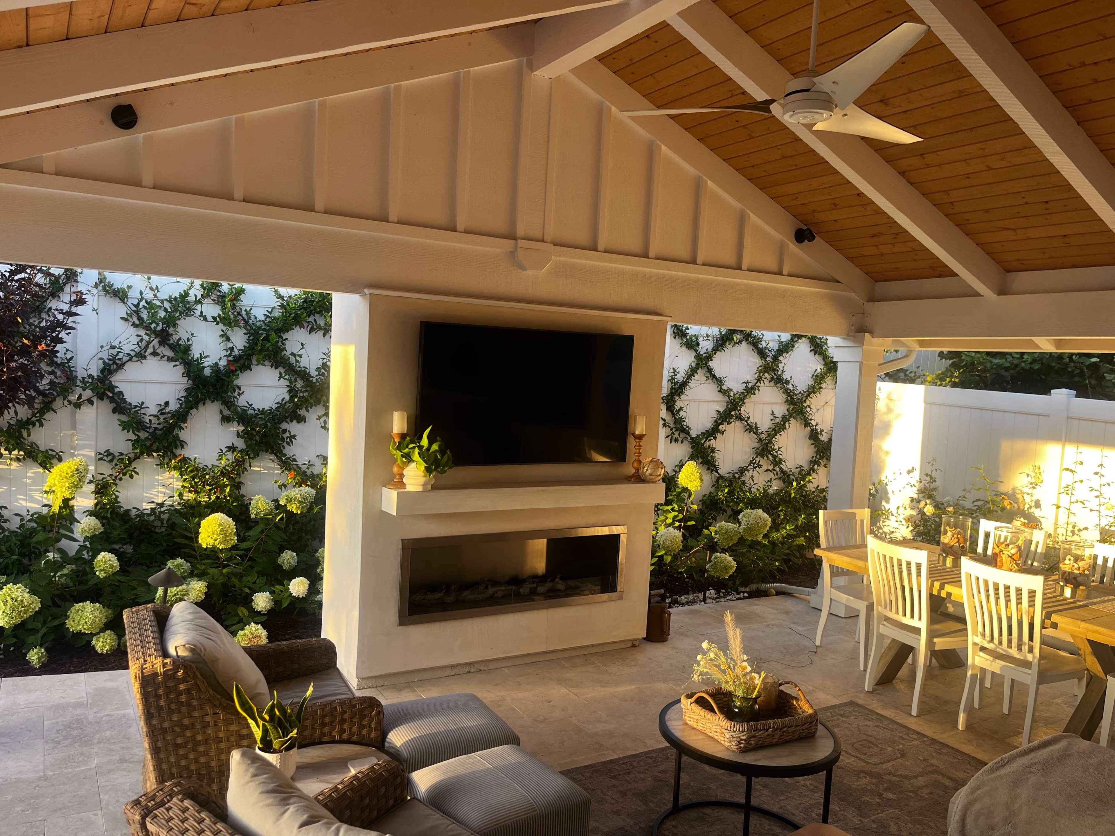 A covered outdoor living area featuring a seating arrangement, a modern fireplace with a television above it, and a dining table, surrounded by landscaped greenery and flowers.