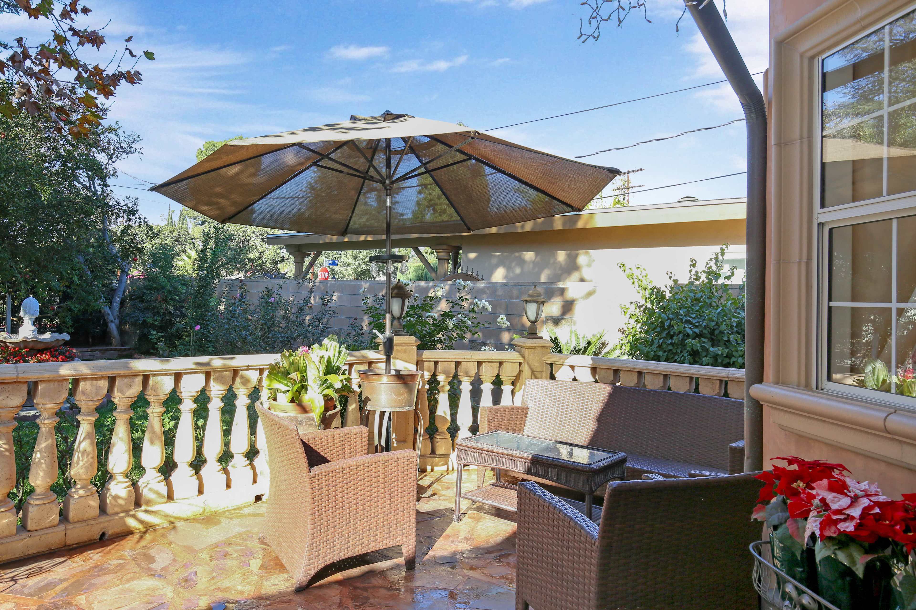 A patio features wicker furniture, a large umbrella, and potted plants surrounded by a stone wall and trees.