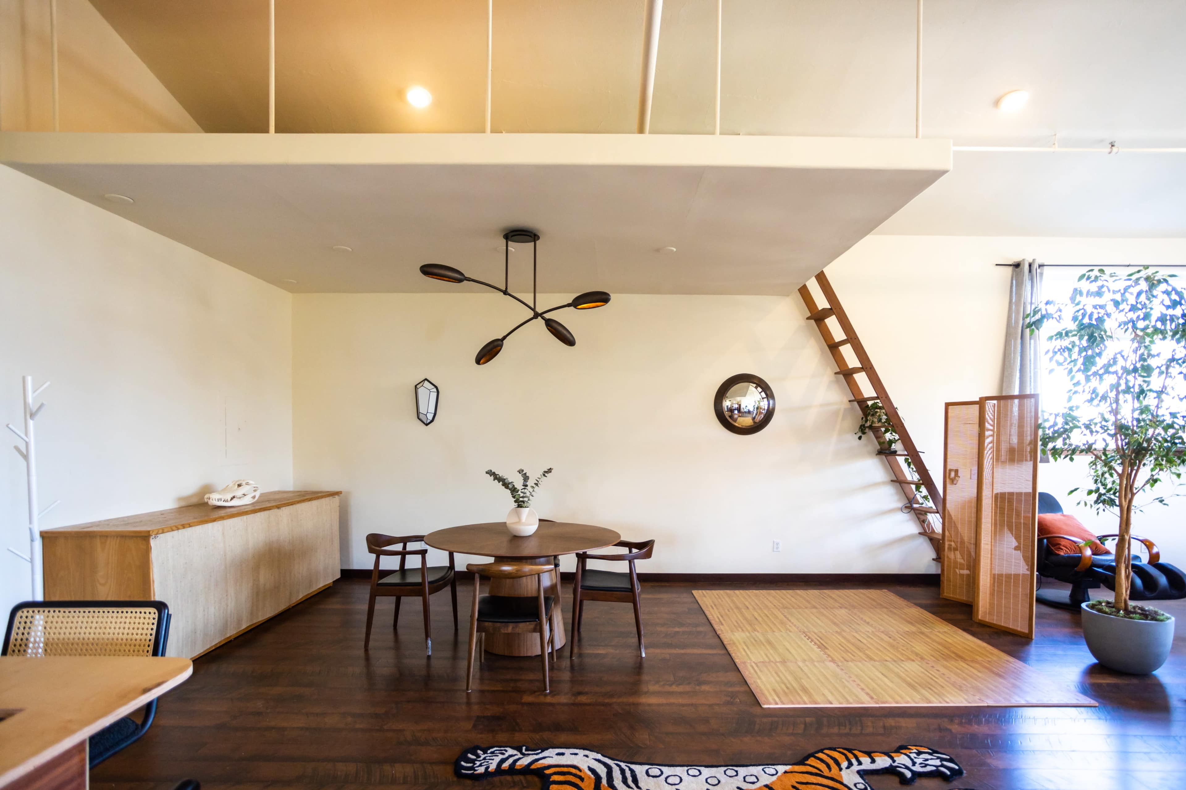 The image shows a minimalist interior featuring a round dining table with chairs, a wall-mounted mirror, a wooden ladder leading to a loft, and plants near the windows.