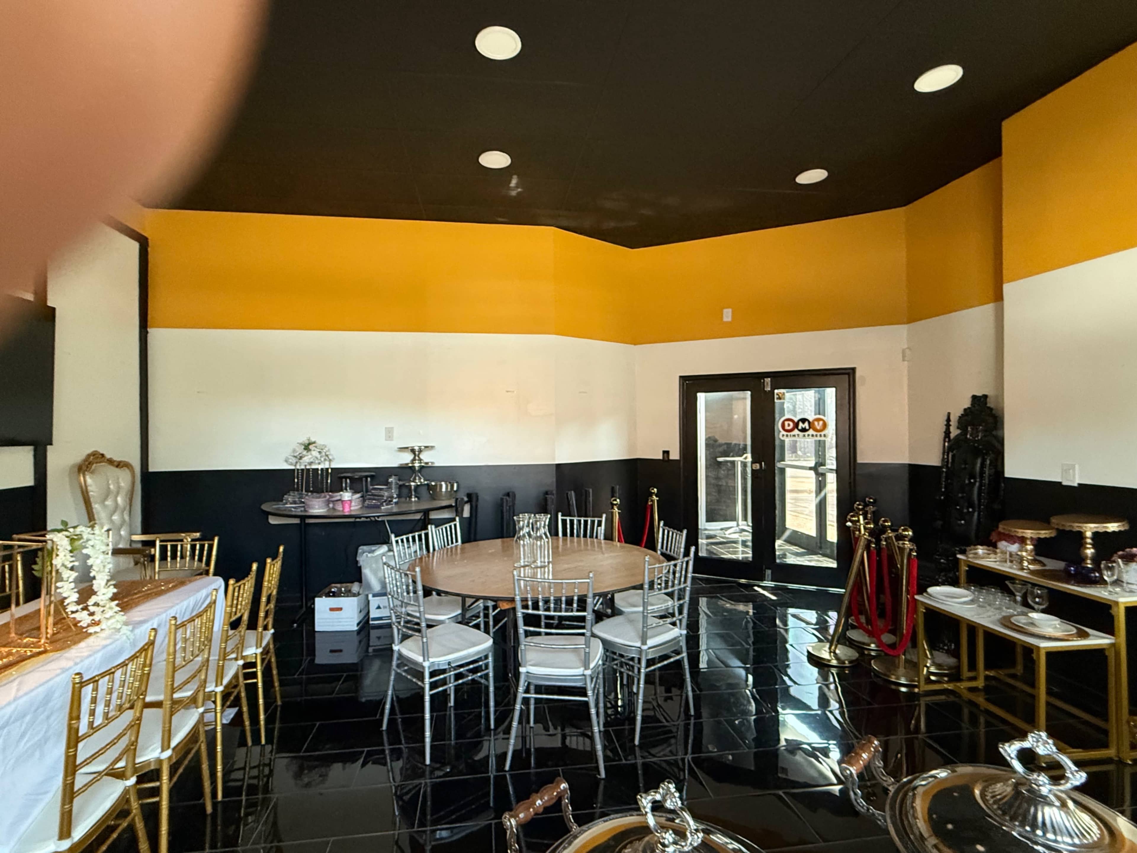 The image shows an interior room with a circular dining table, several chairs, and a buffet setup, featuring black tile flooring and an orange and white color scheme on the walls.