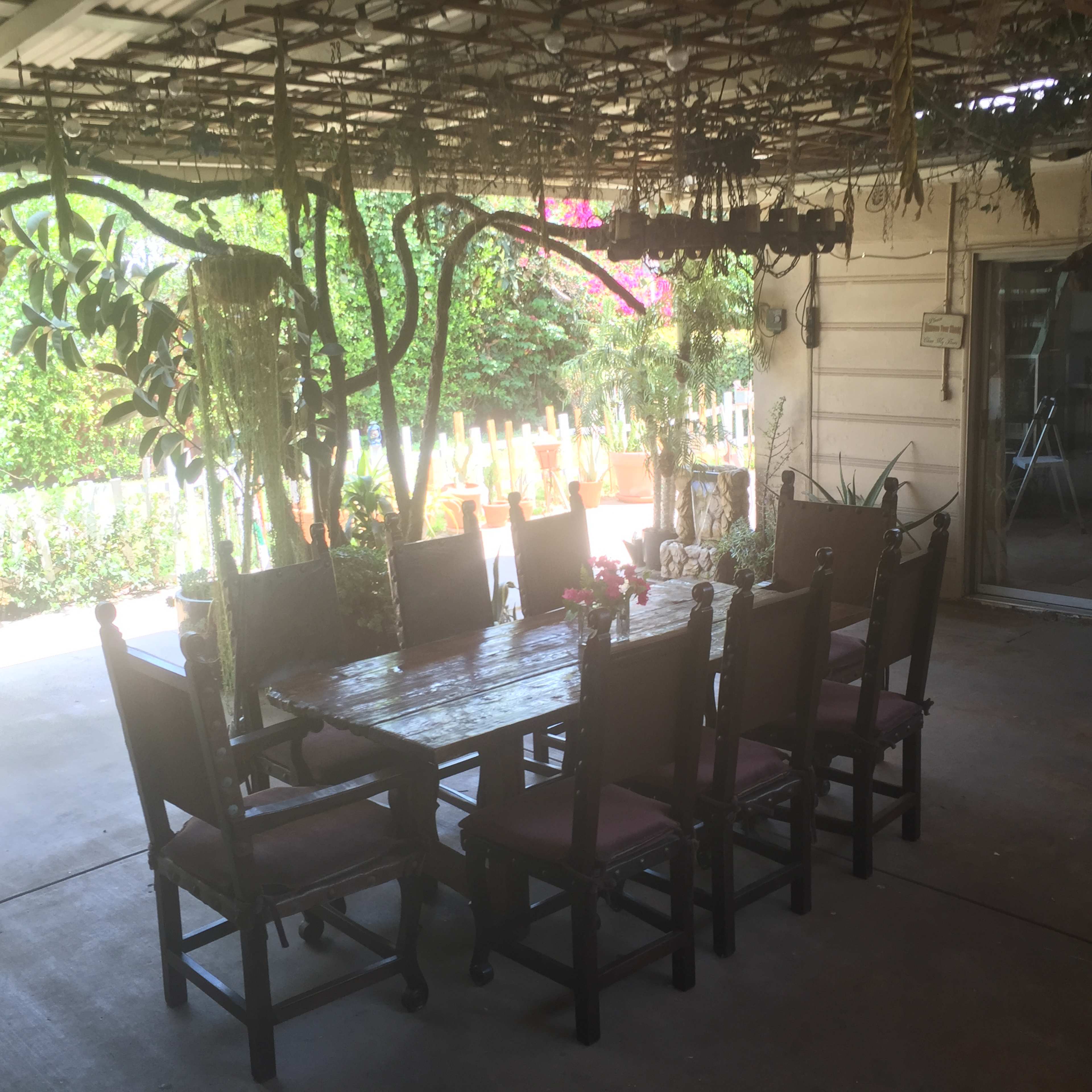 A large wooden dining table surrounded by twelve chairs is set under a shaded patio with hanging plants and a view of a garden.