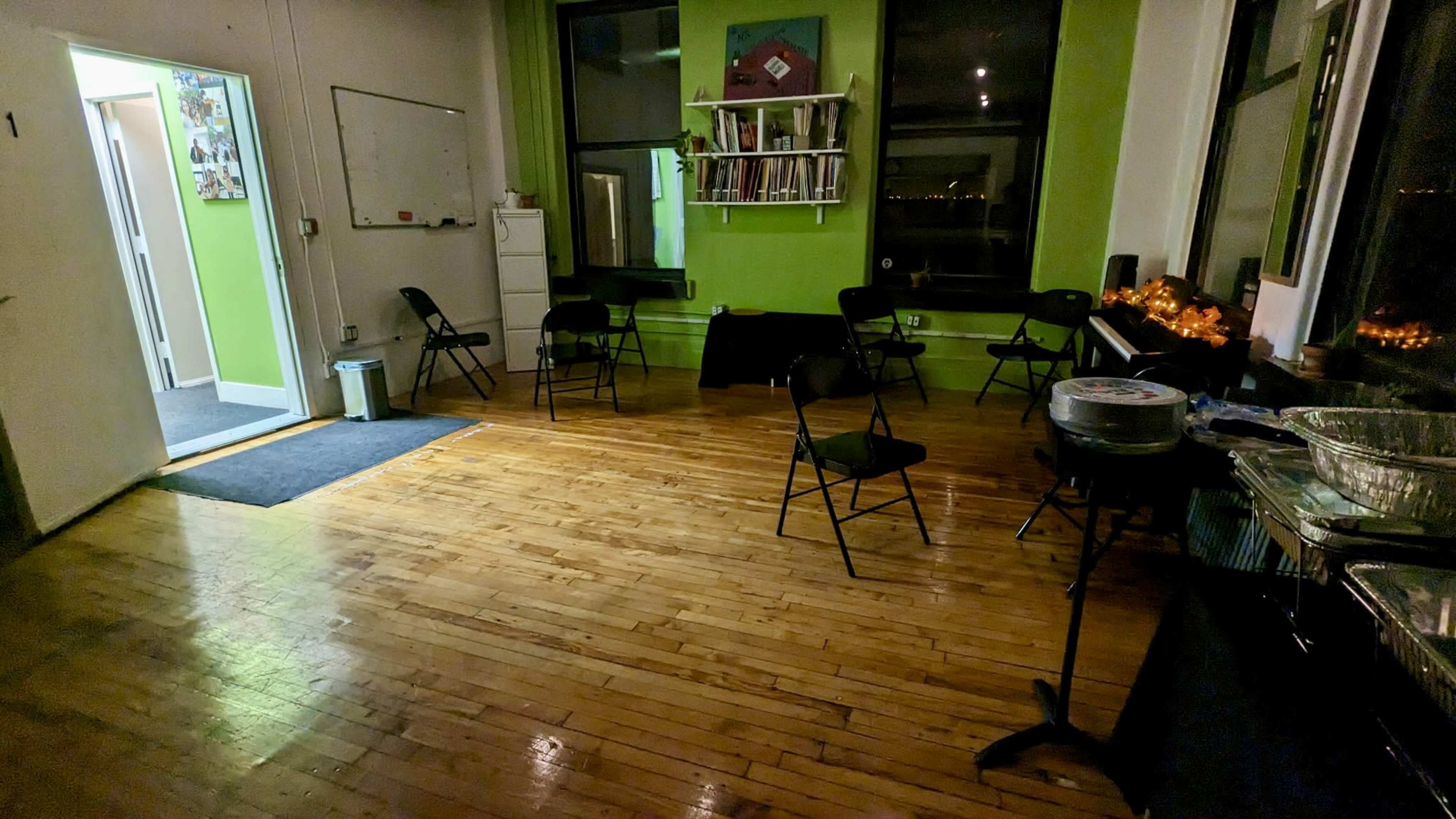 The image shows a sparsely furnished room with wooden floors, several folding chairs arranged in the center, and bright green walls, along with a table holding trays and a piano in one corner.