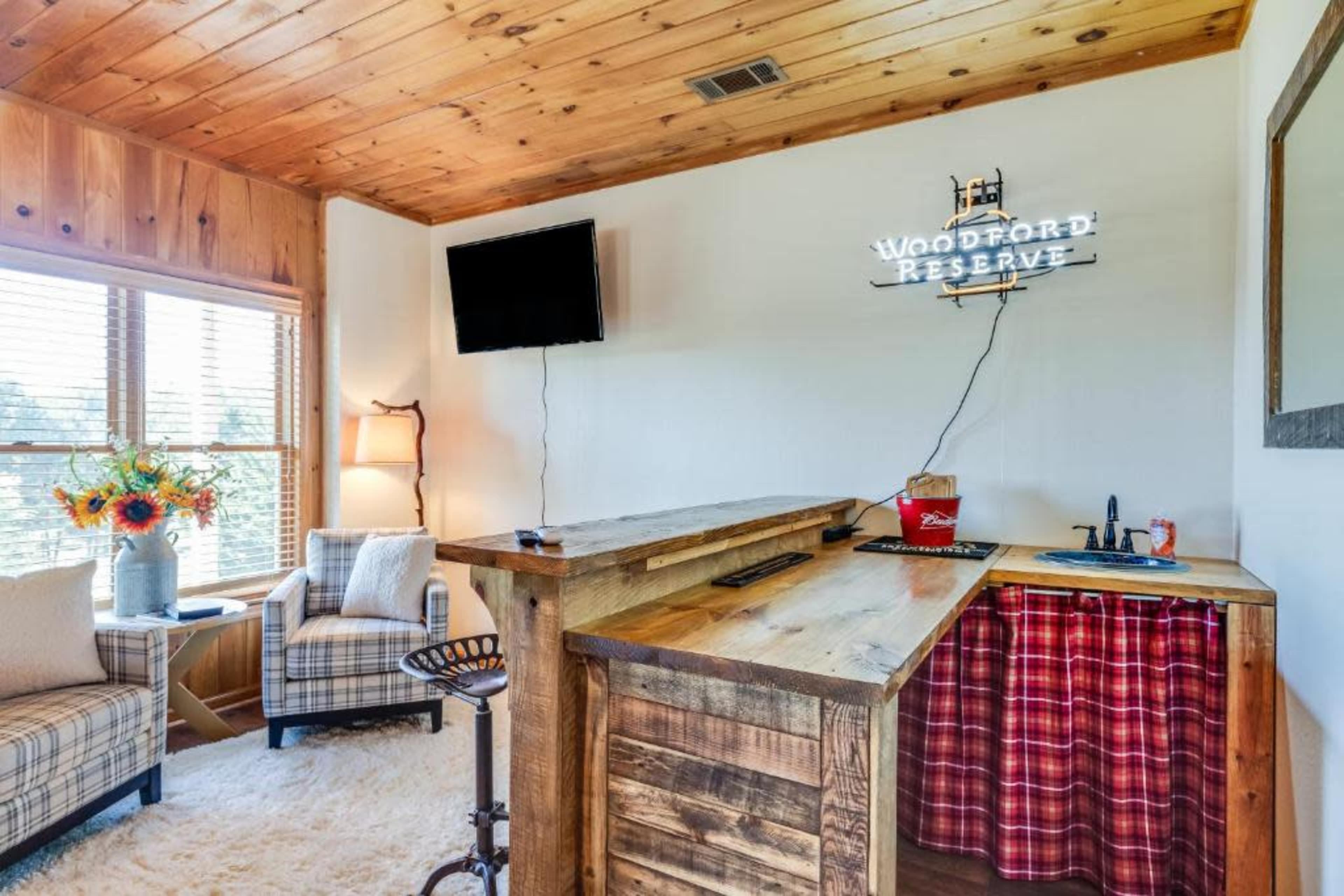 A cozy room featuring a wooden bar with a sink, two armchairs, a flat-screen TV mounted on the wall, and a decorative sign that reads "Woodford Reserve."