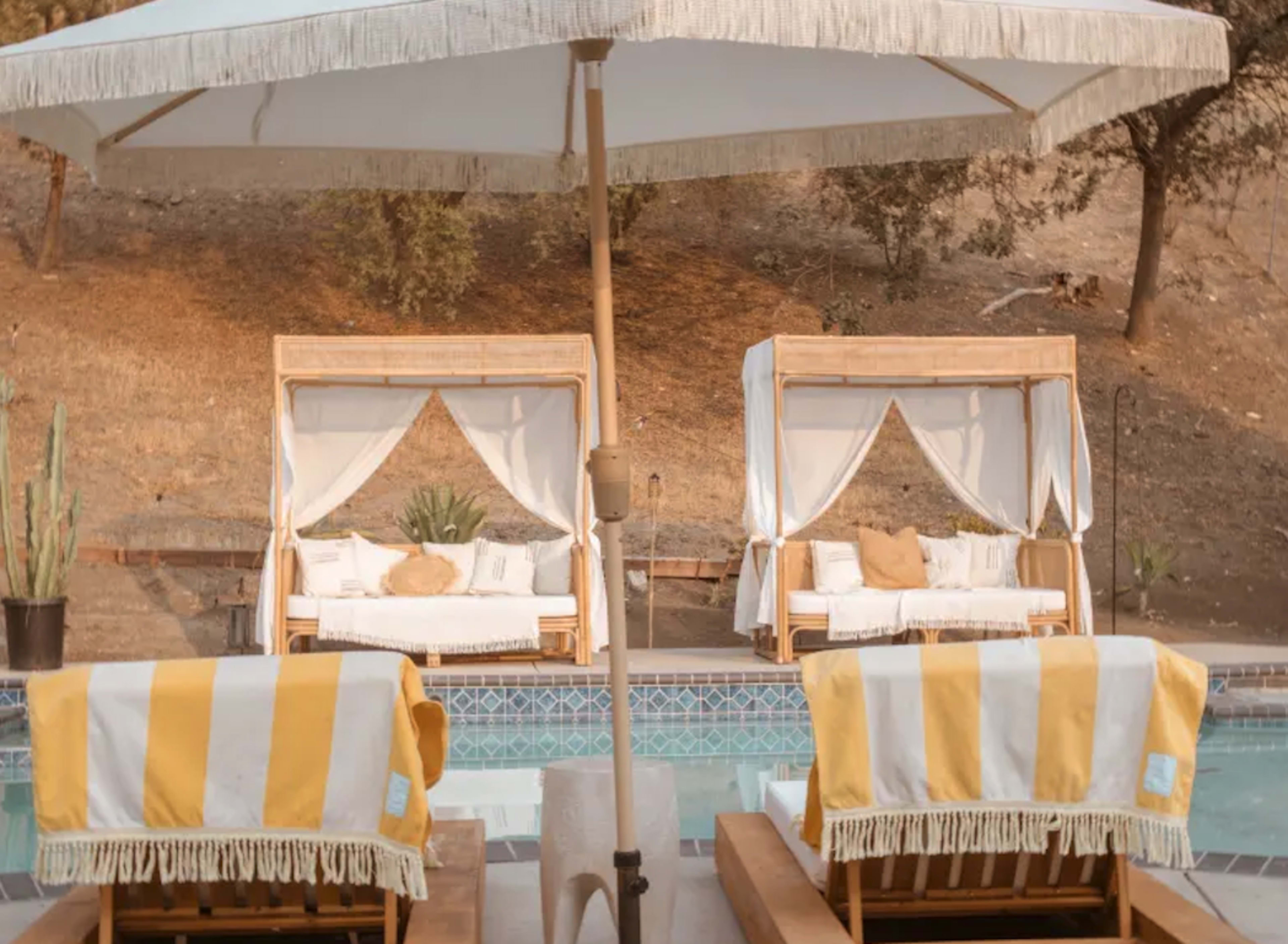 Two cabanas with white drapes and yellow striped lounge chairs beside a swimming pool, framed by a brown hillside and greenery.
