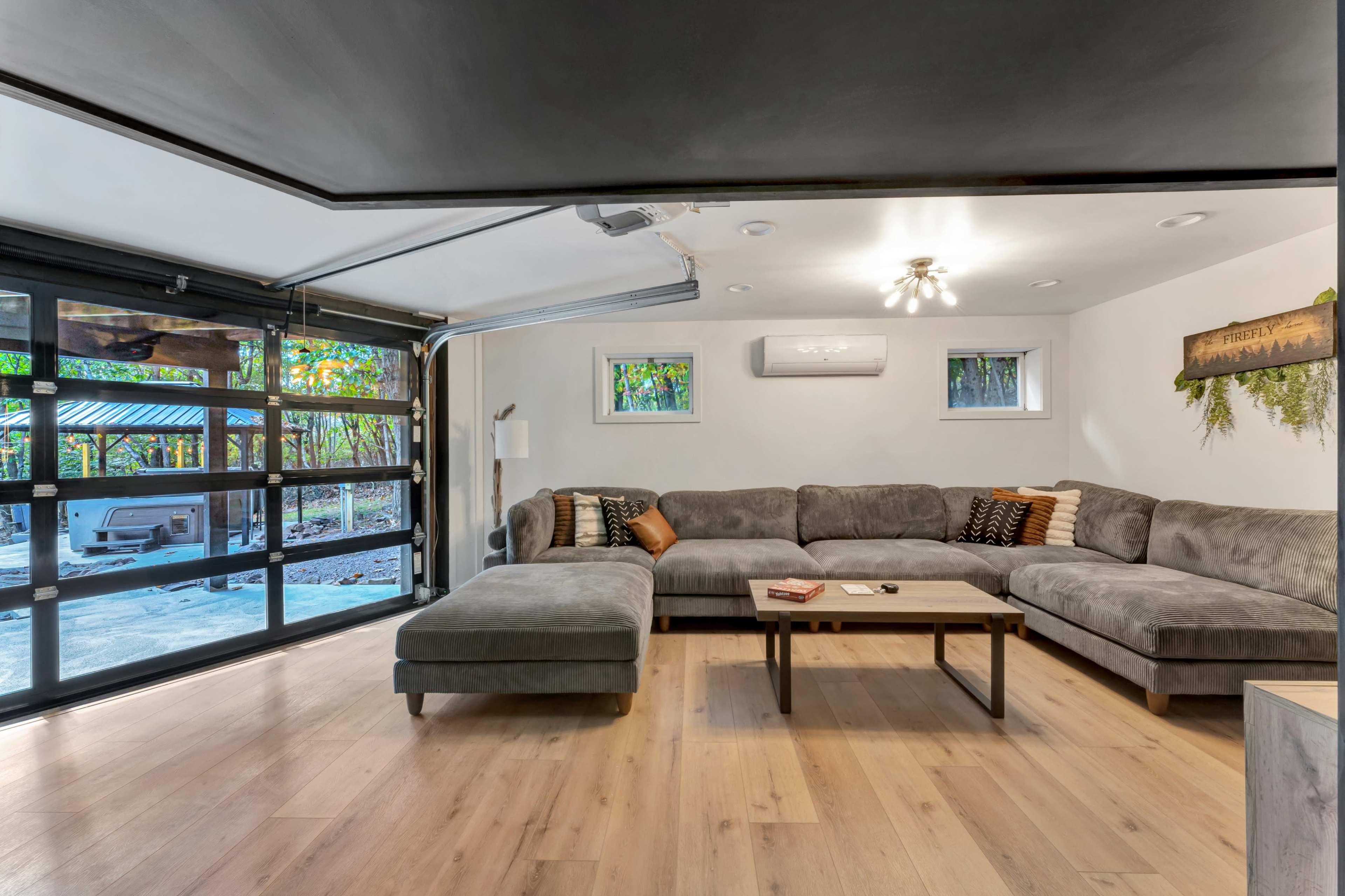 The image shows a modern living room with a large gray sectional sofa, a coffee table, and large windows that provide a view of an outdoor area.