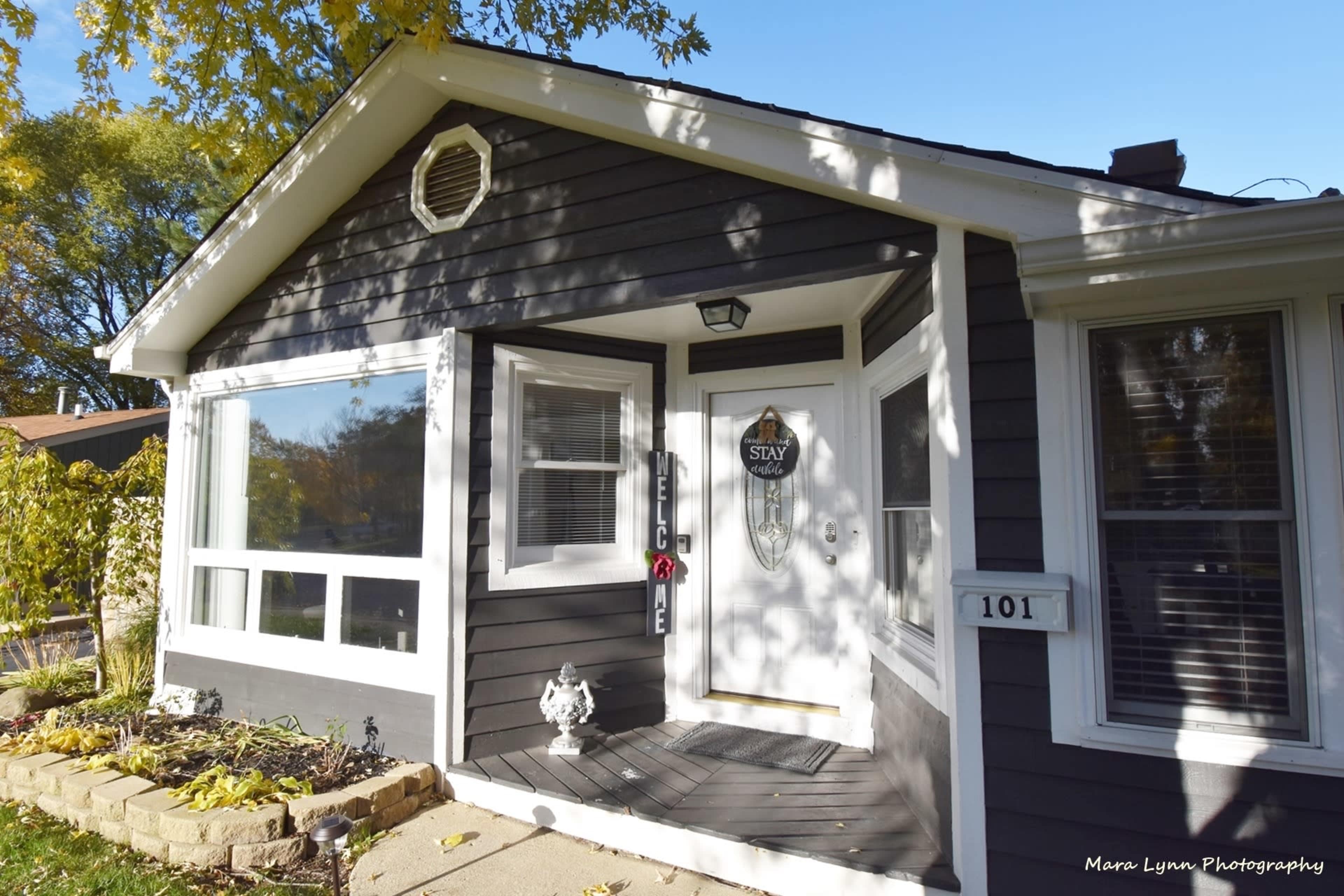 The image shows a modern gray house with a welcoming front porch, featuring a door adorned with a decorative sign and a small garden area.