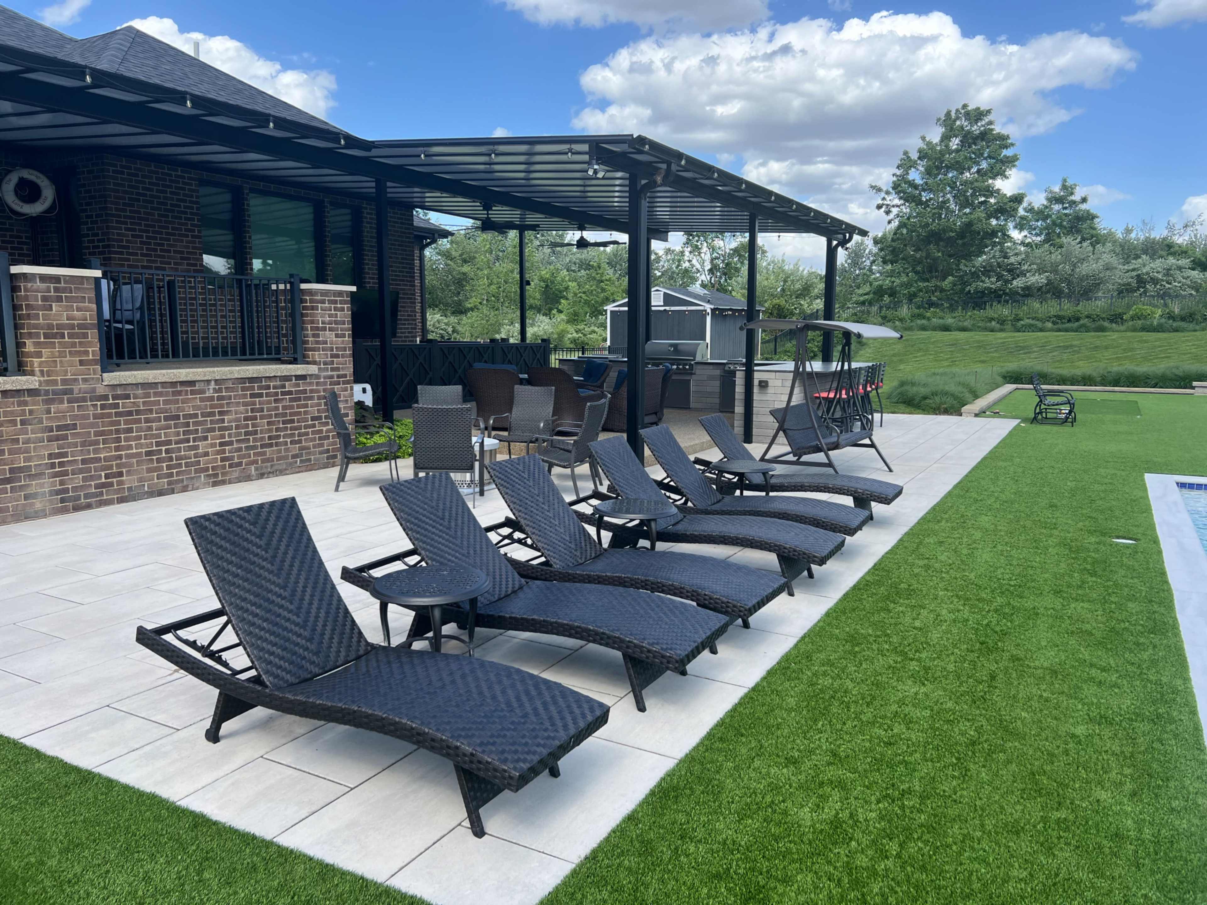 A row of six dark wicker lounge chairs arranged beside a paved pool area with artificial grass and a large house in the background.