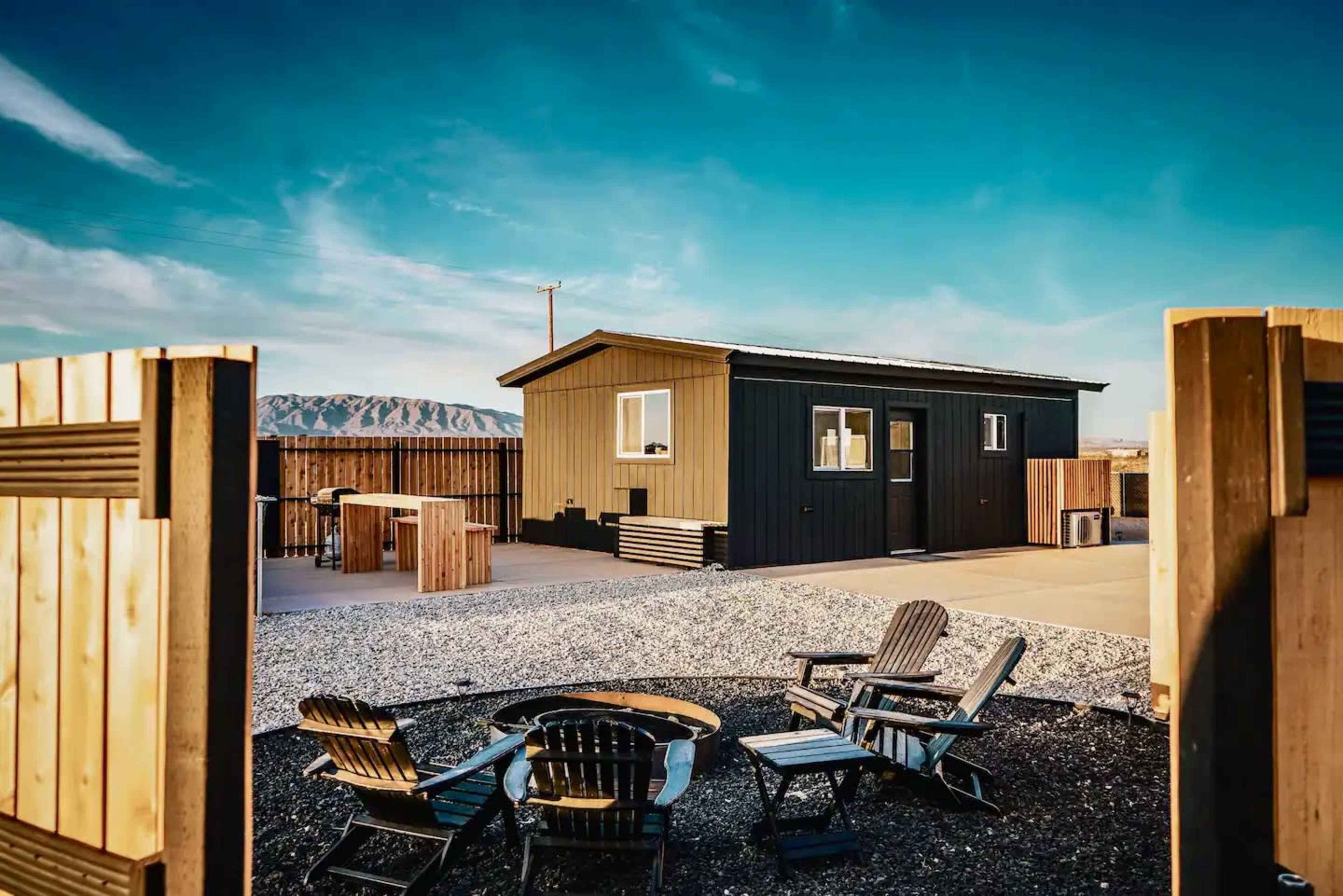 A modern black and yellow house with a gravel yard and a fire pit surrounded by wooden chairs.