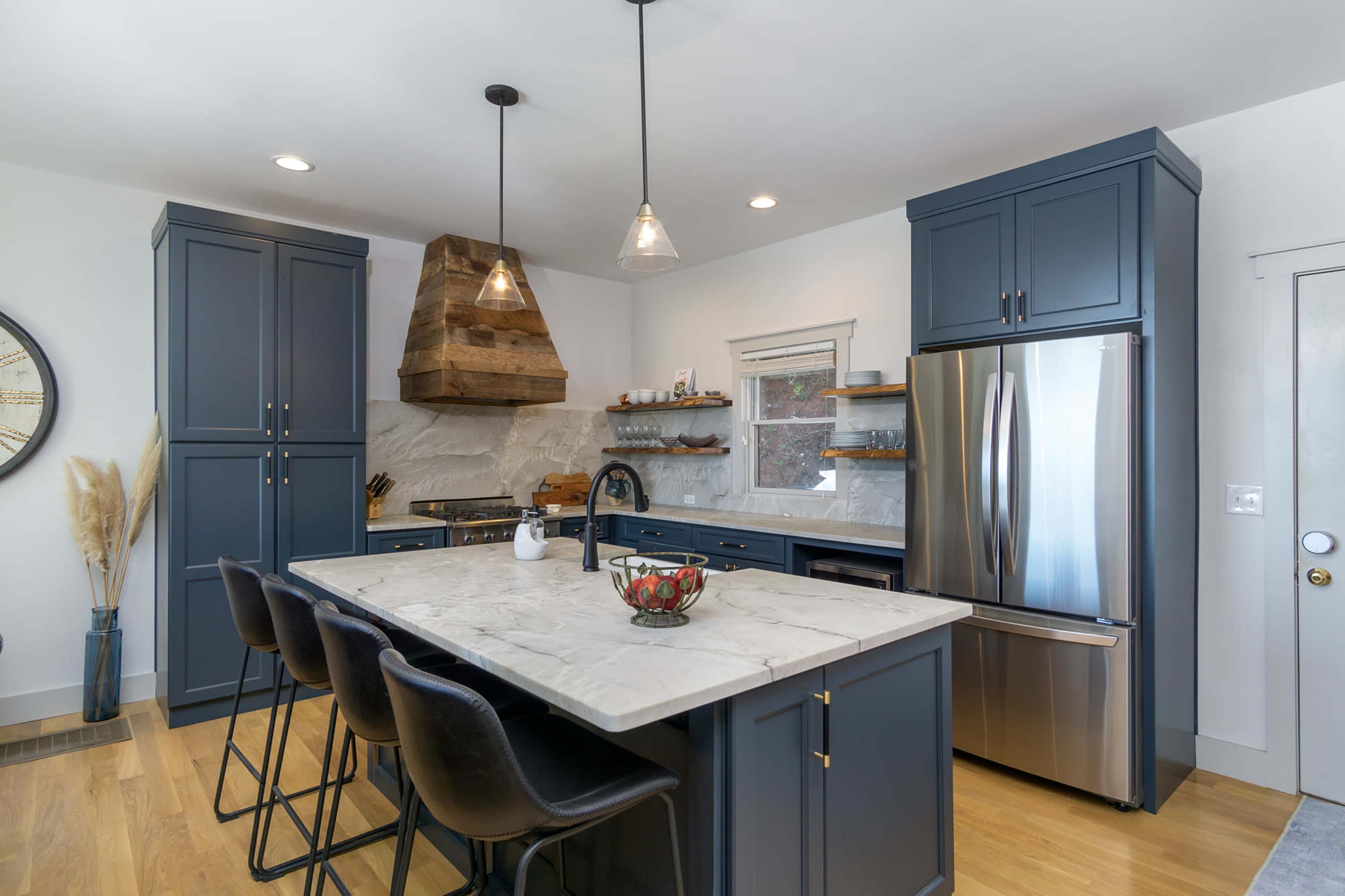 A modern kitchen featuring dark blue cabinets, a large marble island, and a stainless steel refrigerator, all accented by wooden elements and pendant lighting.