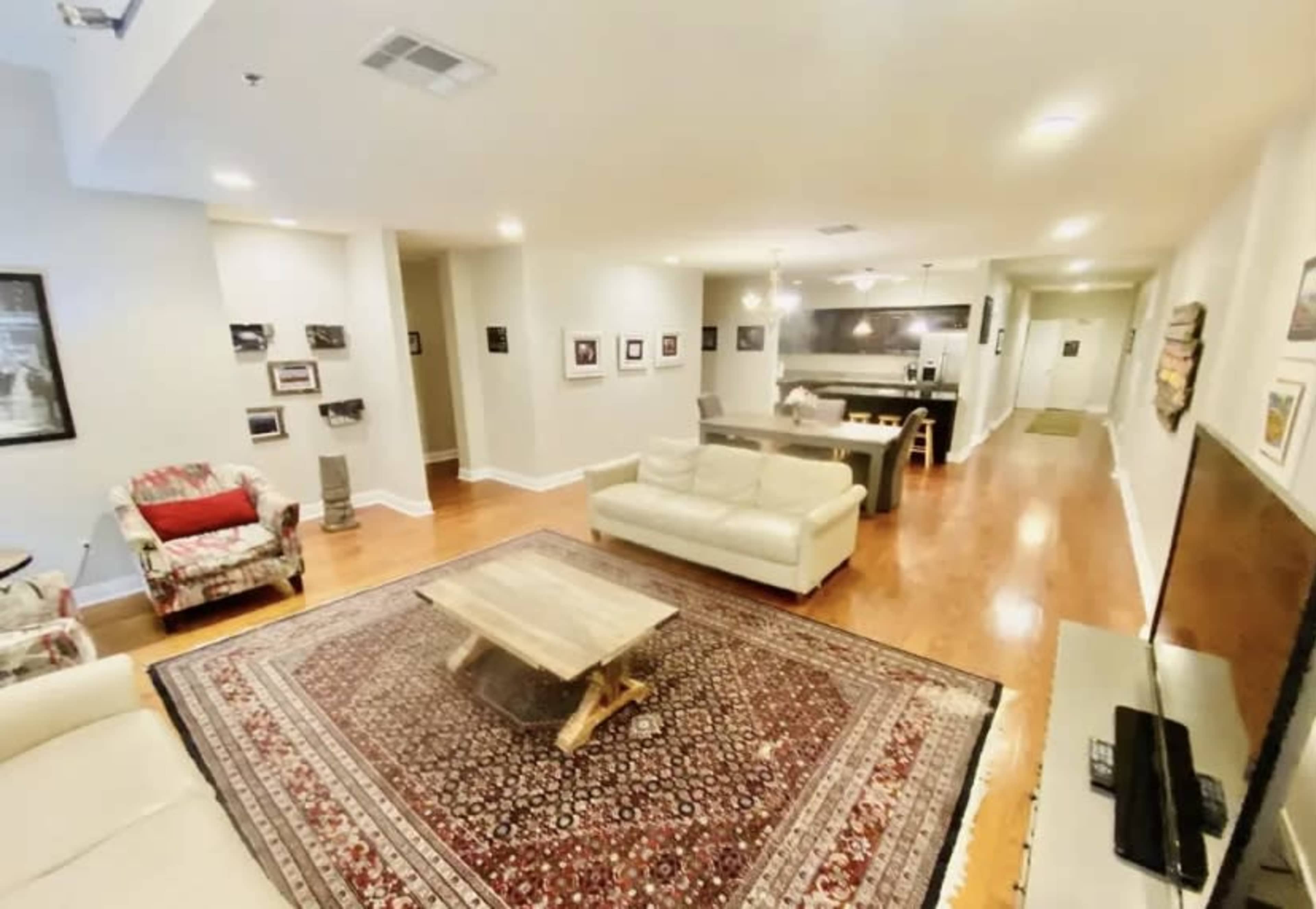 The image shows a spacious living area with two white sofas, a wooden coffee table, a patterned rug, and a wall decorated with framed pictures.
