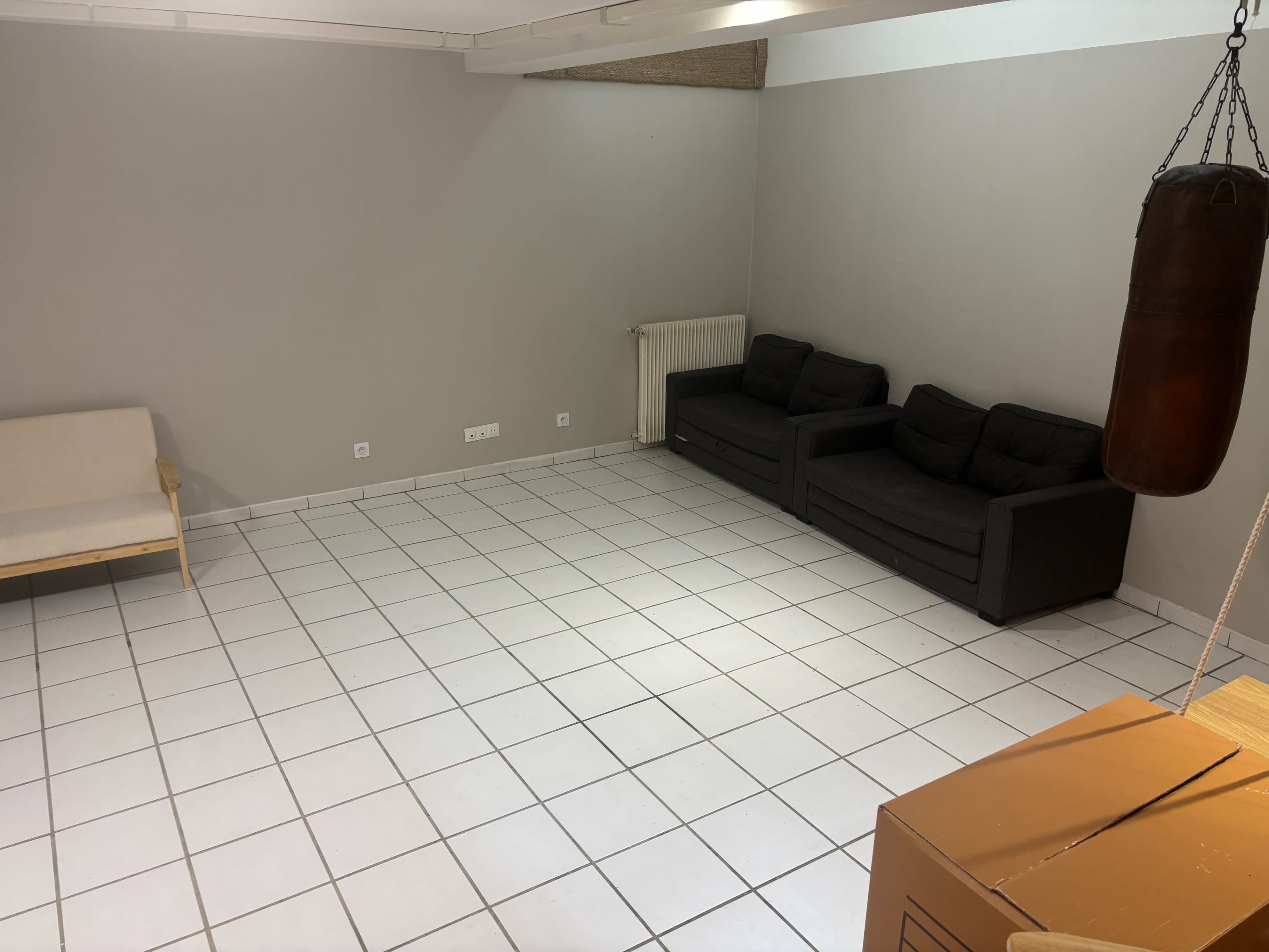 The image shows a sparse room with white tiles, featuring two black sofas, a wooden chair, a punching bag hanging from the ceiling, and a radiator against the wall.