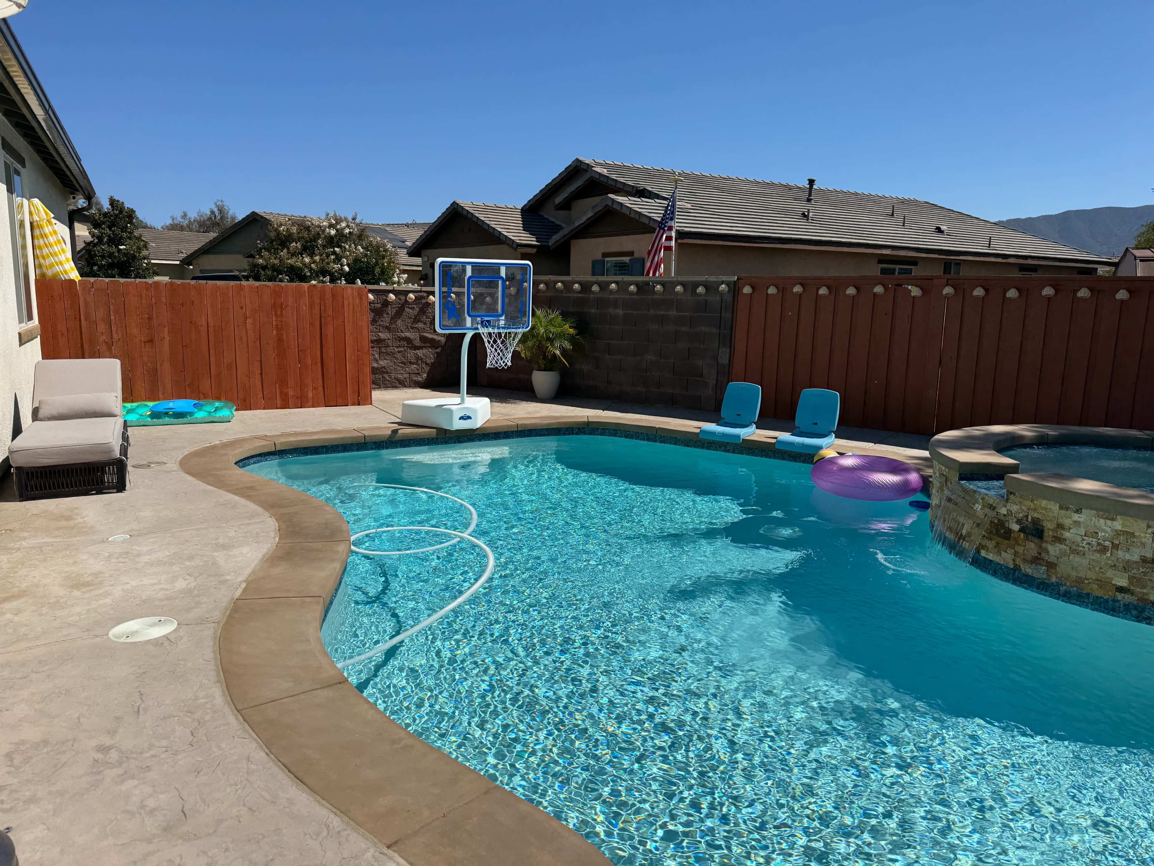 The image shows a residential backyard pool area featuring a clear blue swimming pool, a basketball hoop, lounge chairs, and colorful pool floats.