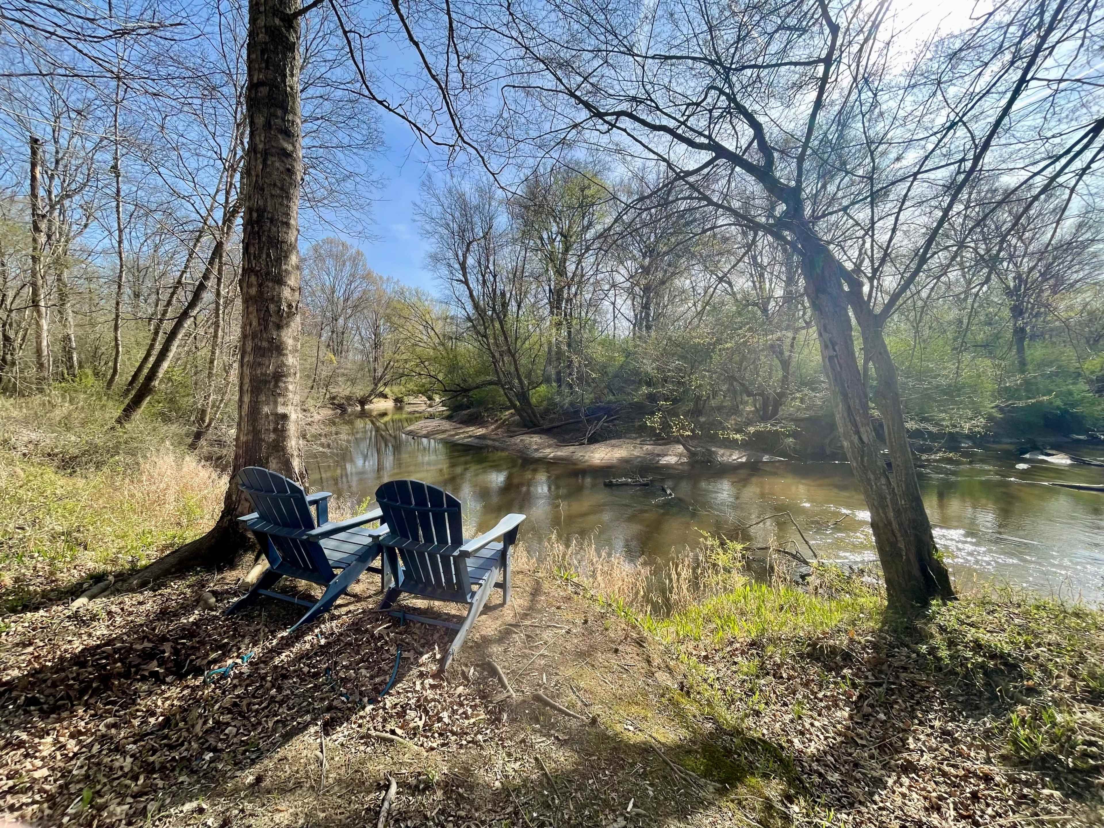 Two blue adirondack chairs are positioned by a calm stream surrounded by trees and greenery.