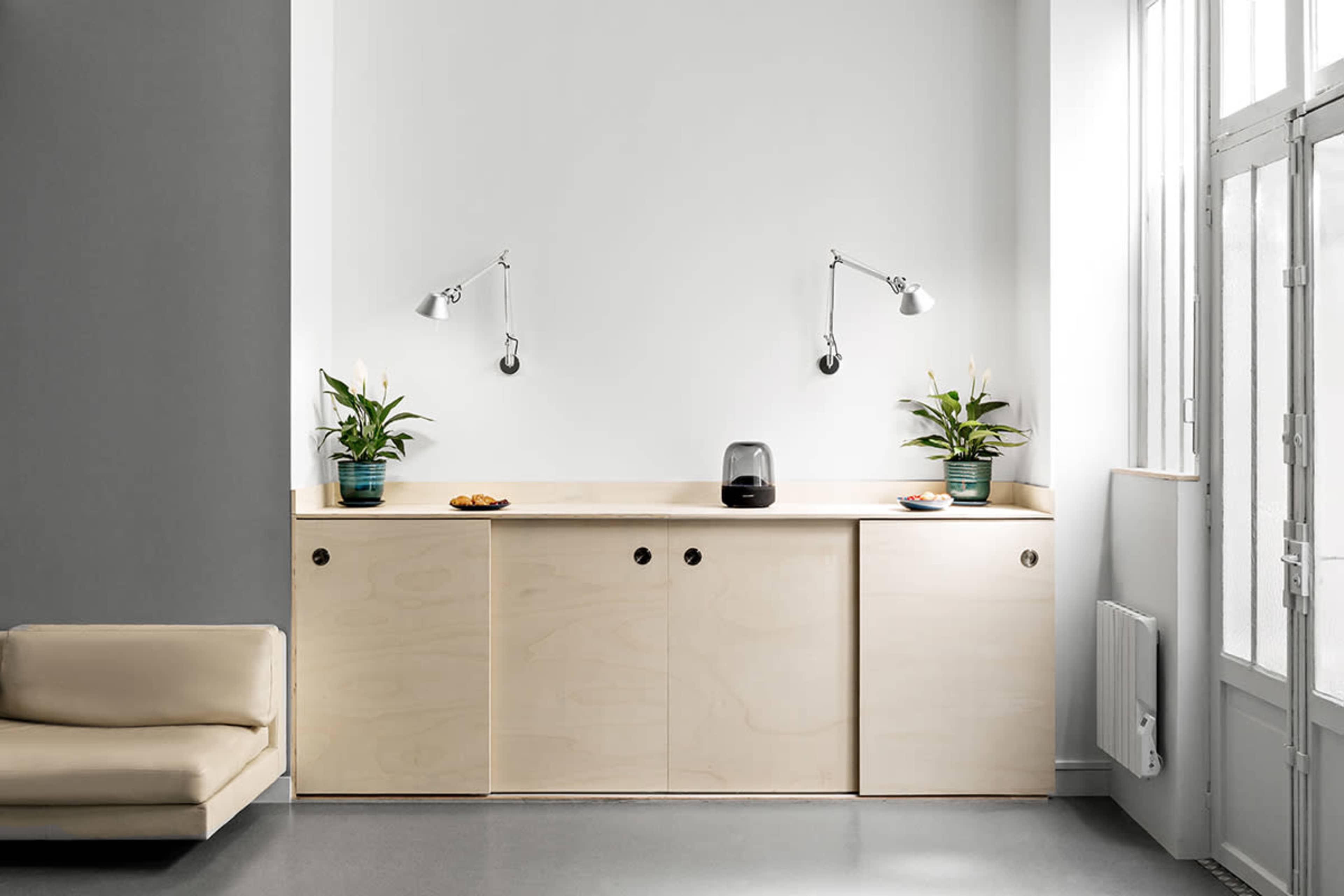 A minimalist interior with a light wood cabinetry unit, two tabletop plants, and wall-mounted lamps, set against a simple white wall.