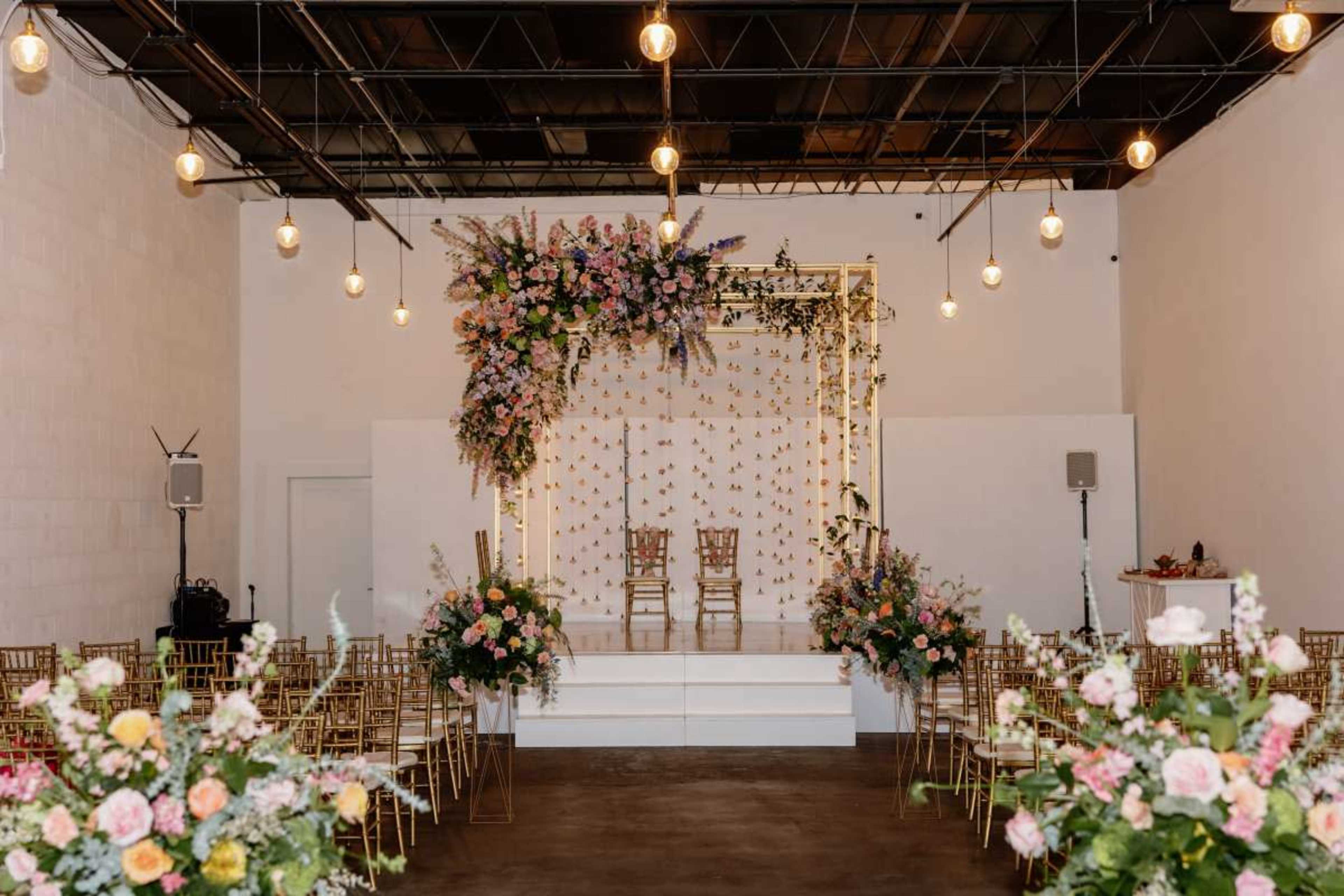 A wedding ceremony setup features a floral archway, two chairs on a stage, and rows of gold chairs adorned with flowers in a well-lit indoor venue.