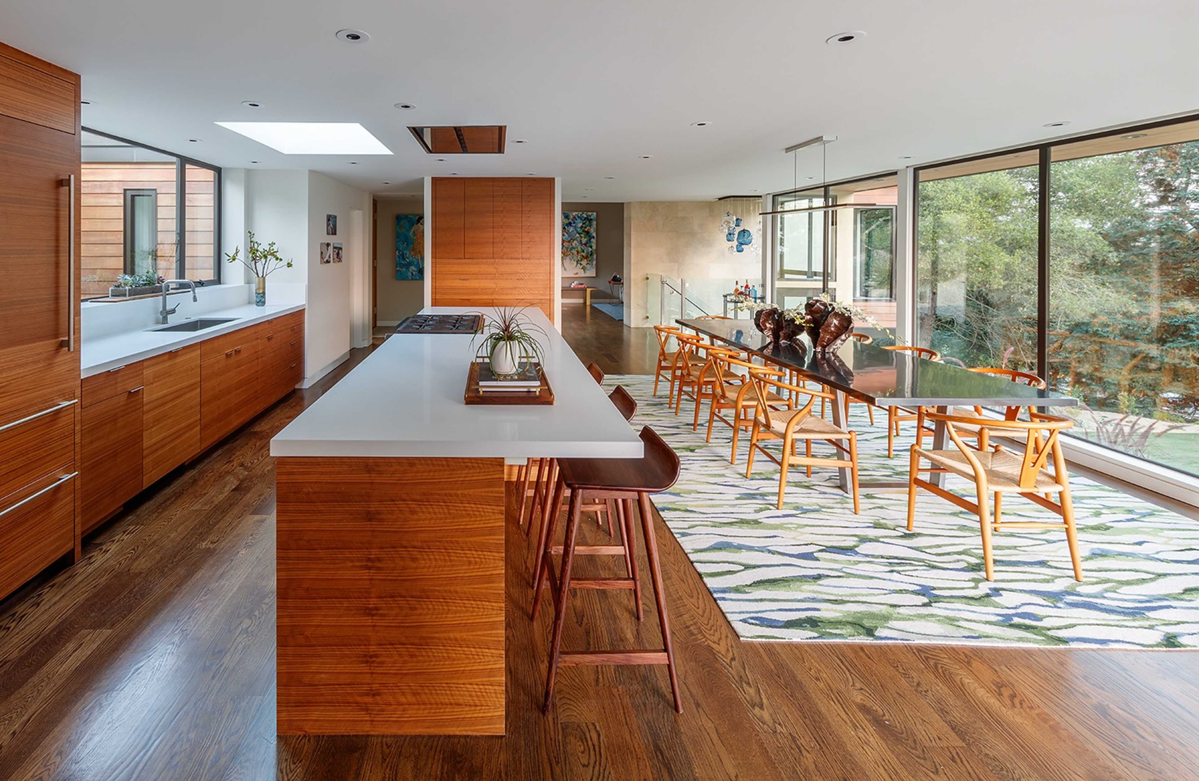 A modern kitchen features wooden cabinetry, a central island with bar stools, and a dining area lined with large windows overlooking a garden.