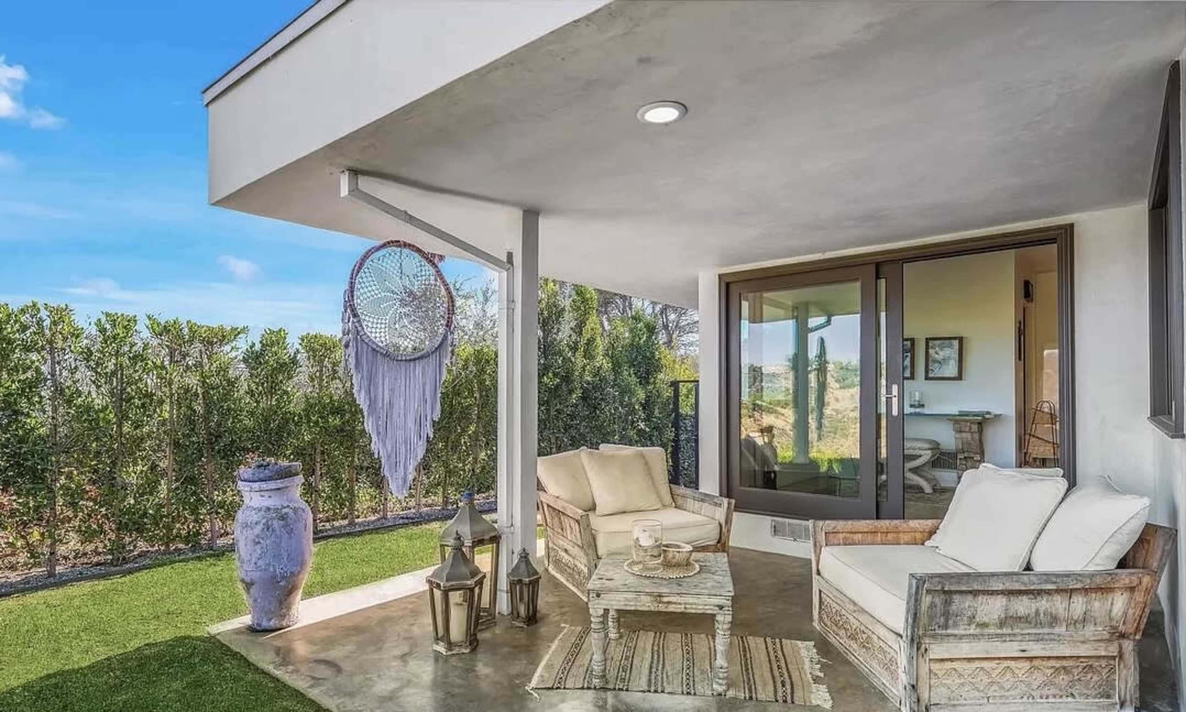 A small, modern porch area features two chairs and a table, with a hanging dreamcatcher and a view of greenery in the background.