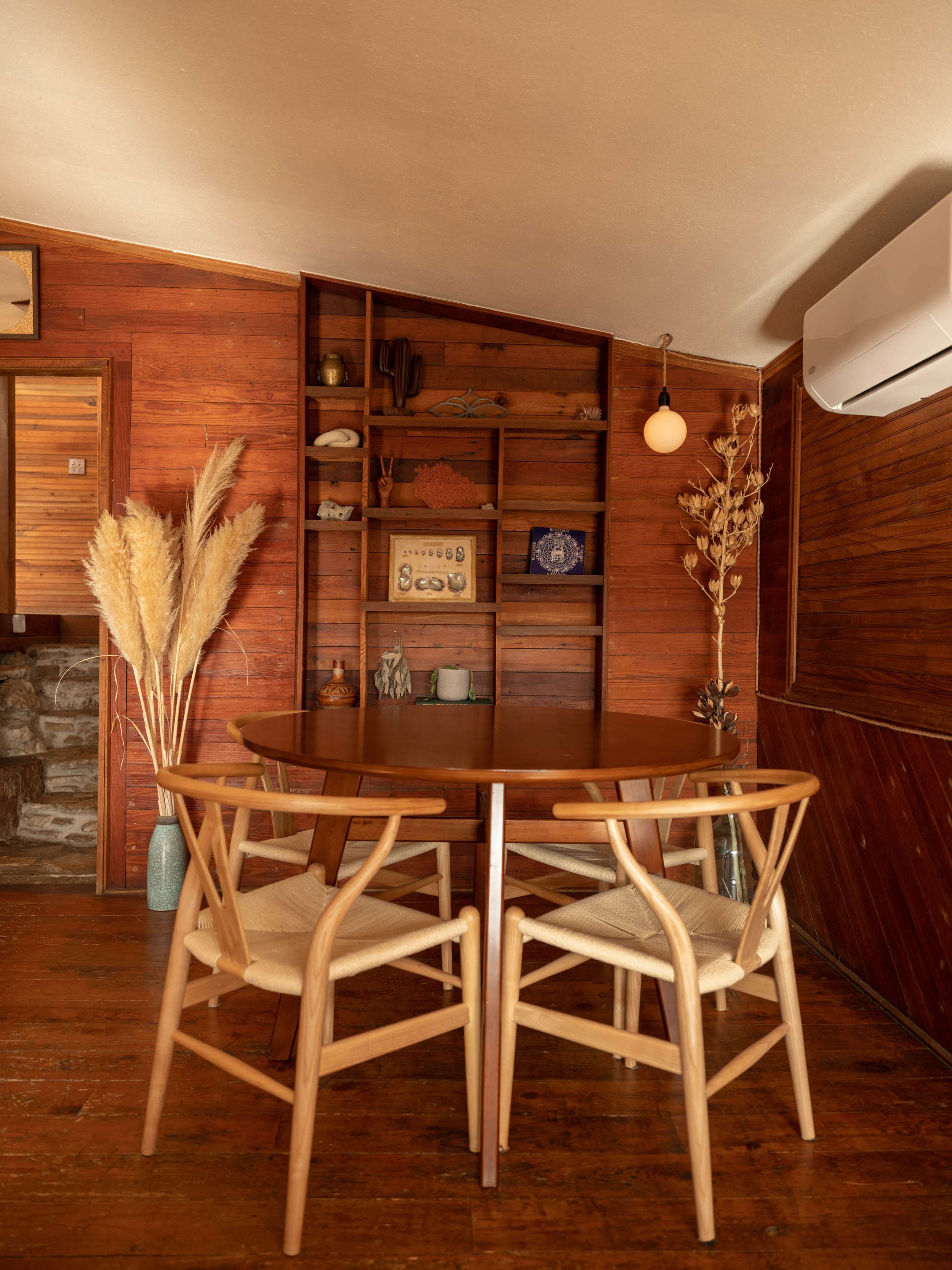 A wooden dining table with two chairs is placed against a rustic wood-paneled wall featuring shelves with decorative items and plants.