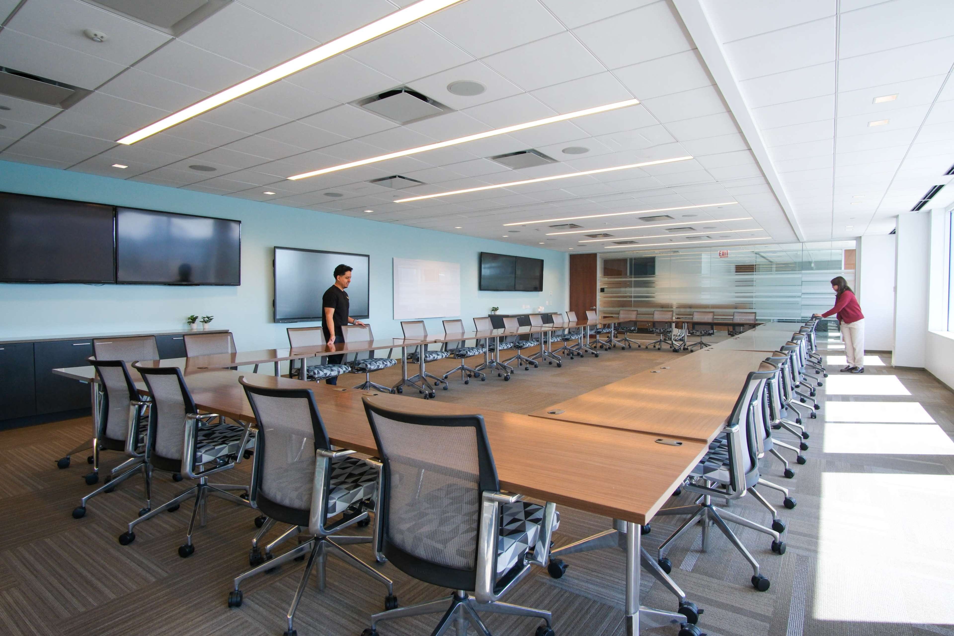 A spacious, modern conference room features a long wooden table with rolling chairs, large screens mounted on the walls, and two people preparing the space.