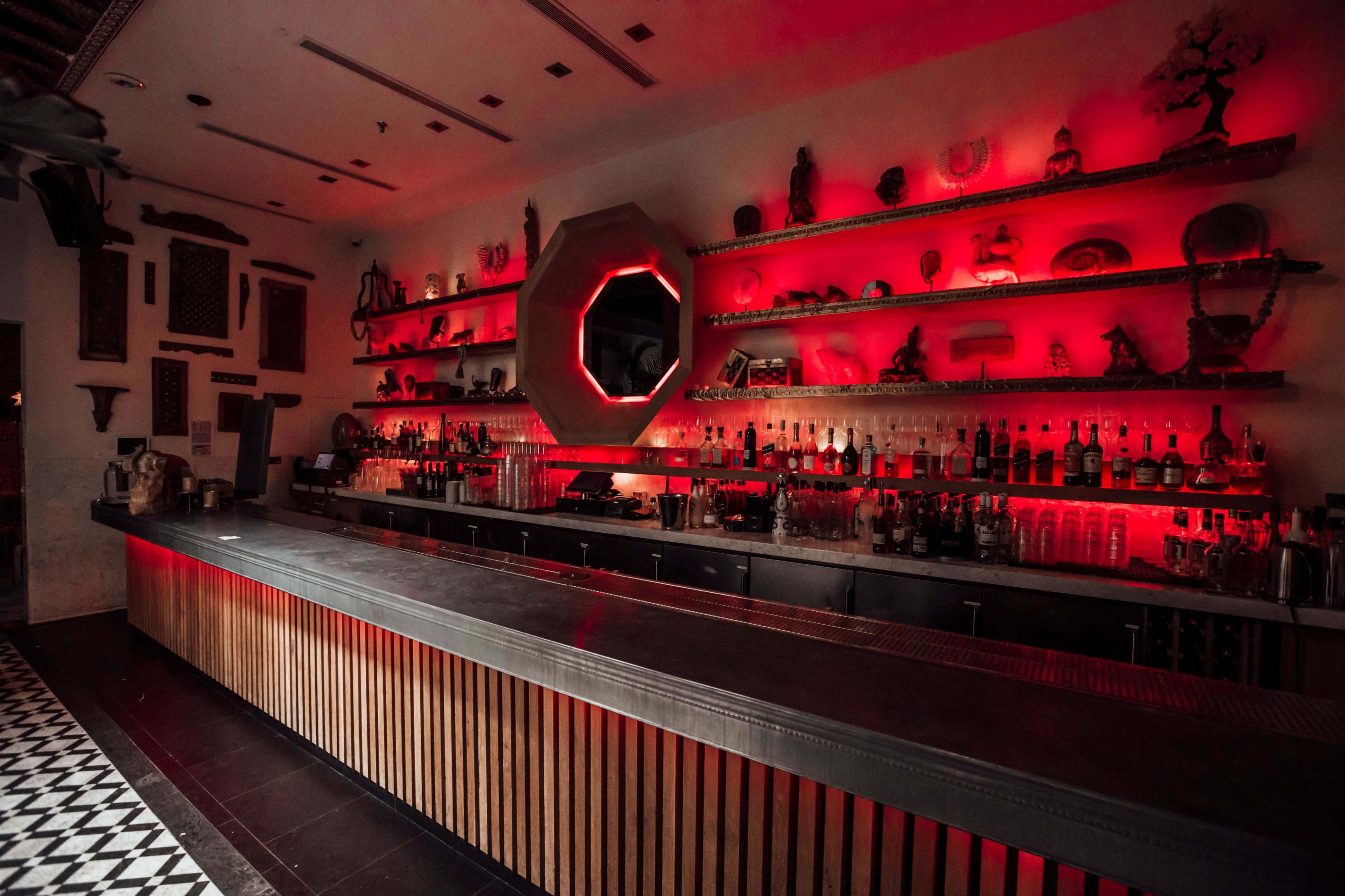 A dimly lit bar with red lighting, showcasing a wooden counter and shelves filled with various bottles and decorative items.