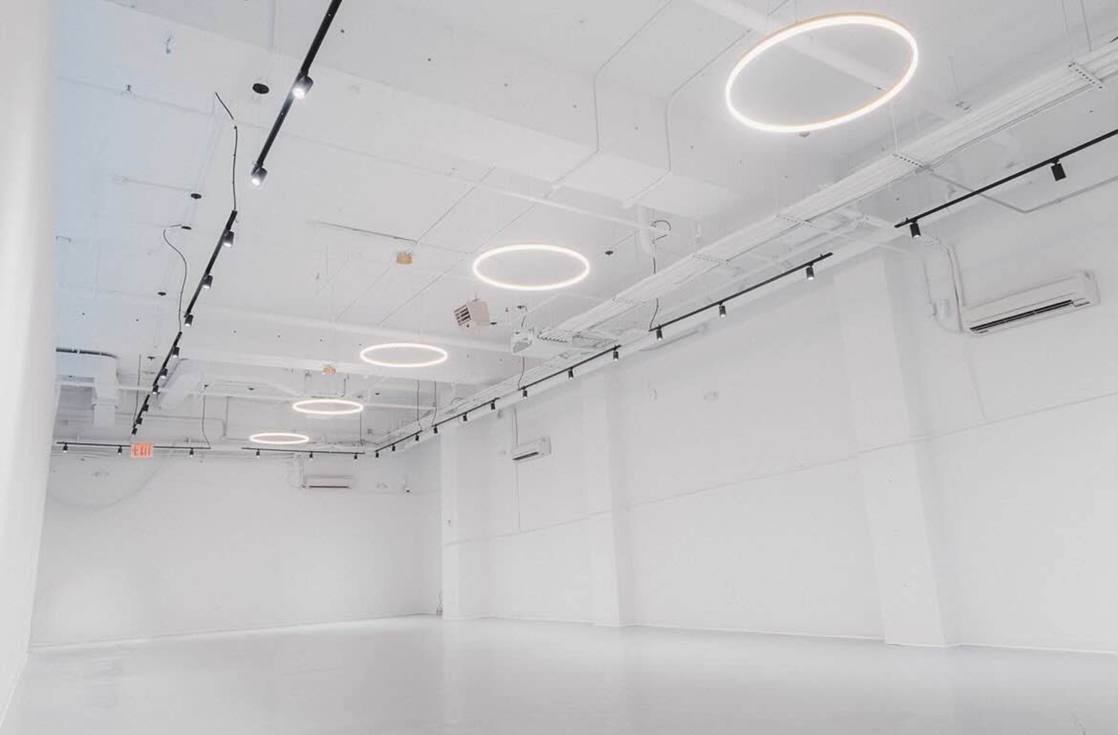 The image shows a spacious, empty room with white walls and ceiling, featuring circular lights suspended from the ceiling.